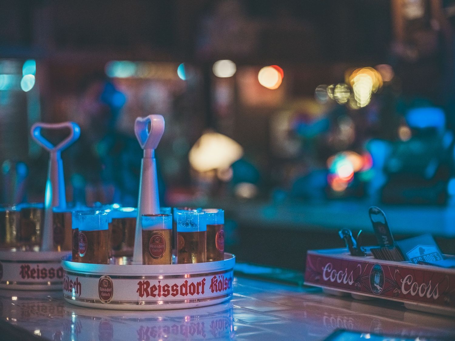 Reissdorf Kölsch beer tray with glasses and Coors ashtray on a bar counter with colorful bokeh lights.