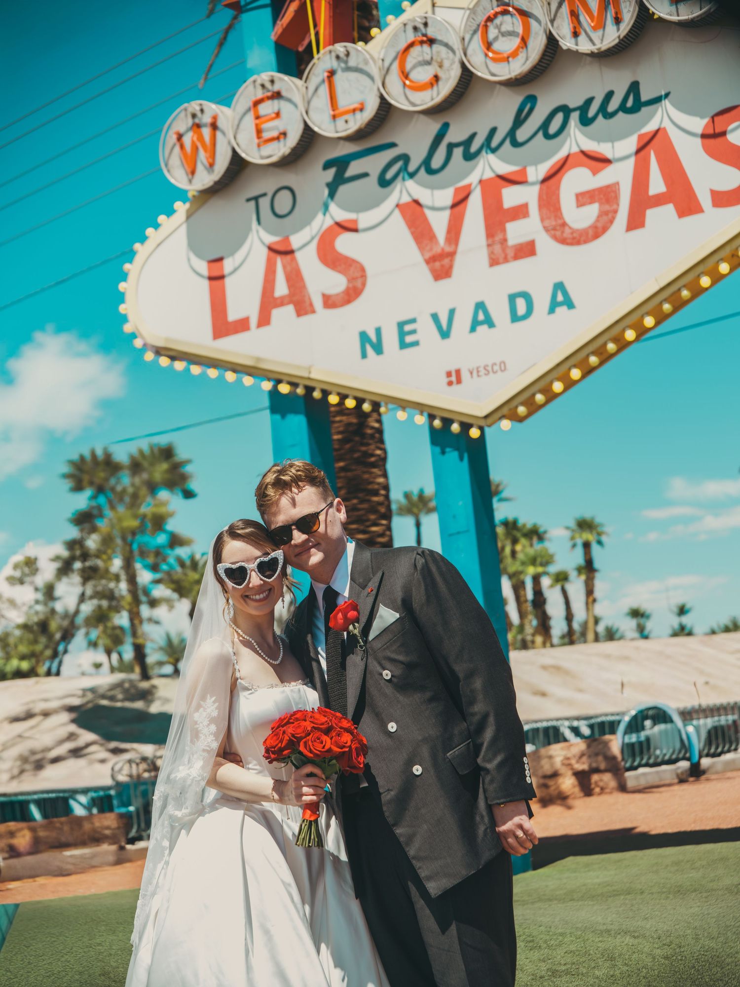 Newlywed couple in wedding attire and heart sunglasses posing in front of the Welcome to Fabulous Las Vegas Nevada sign.