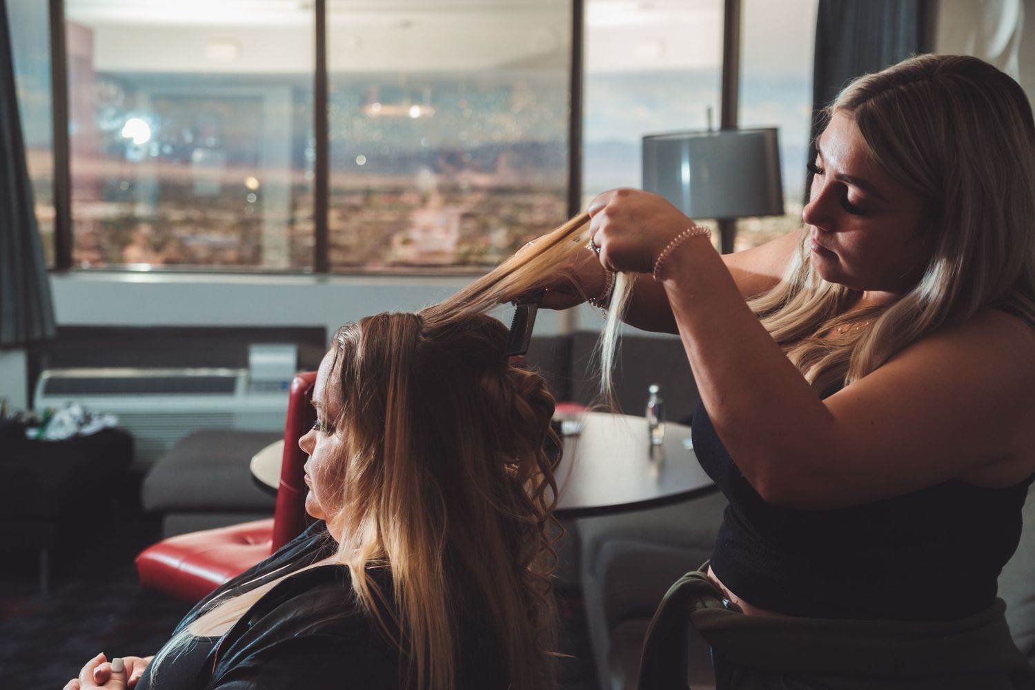 A hairstylist curling long brown hair with a flat iron in a modern hotel room with city views.