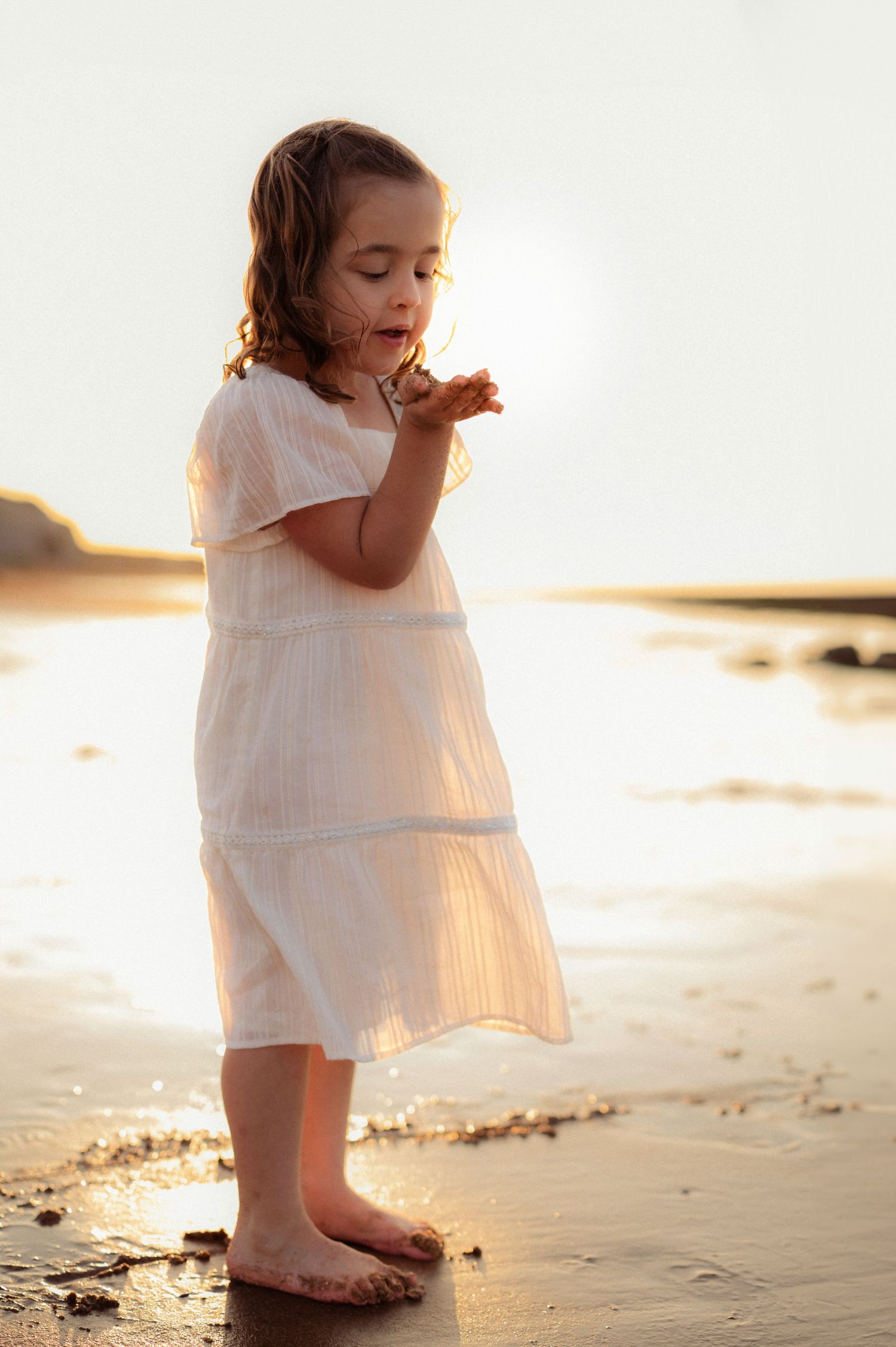 Little girl in white dress standing barefoot on wet sand blowing something from her hand at sunset.