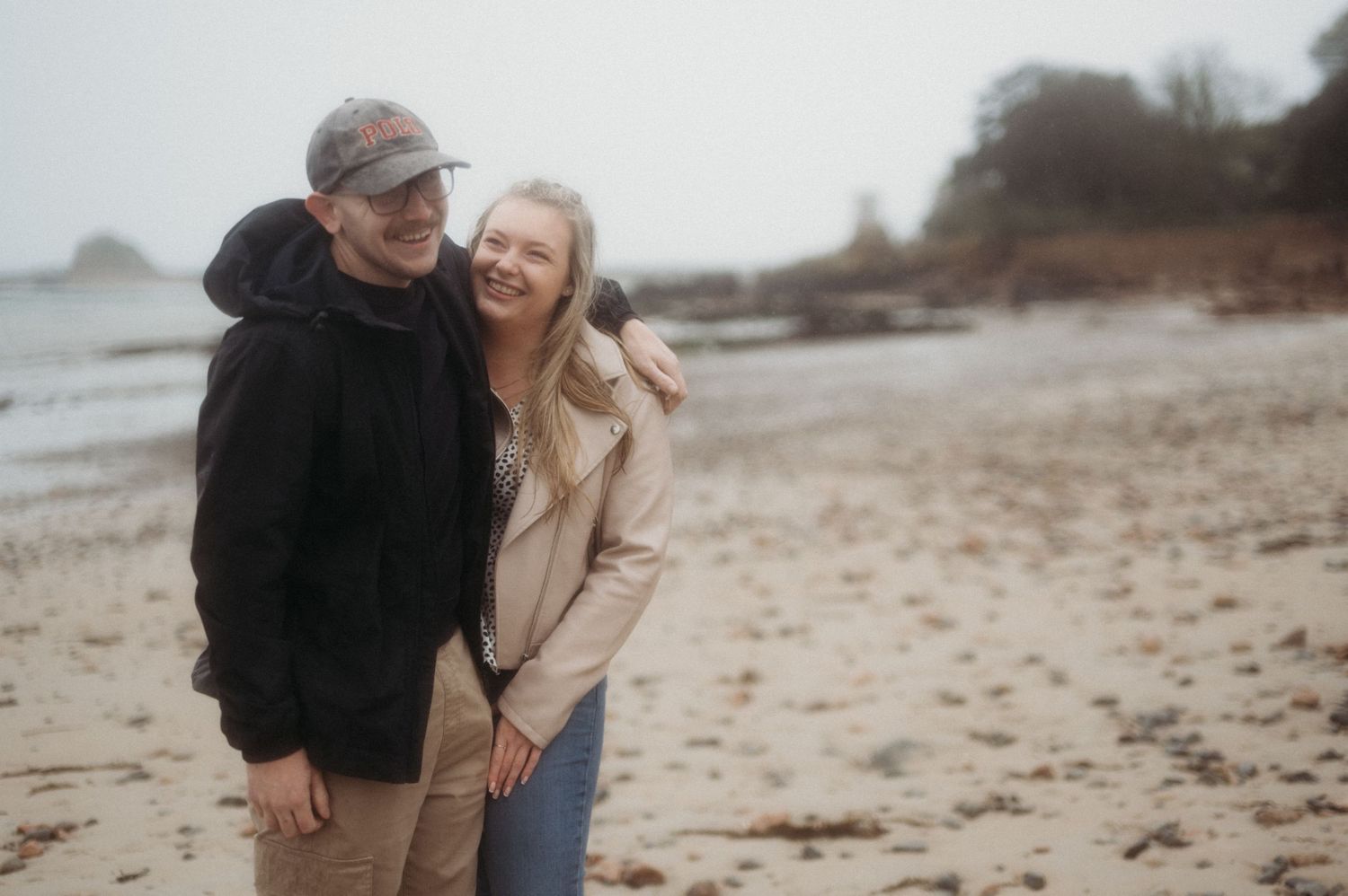 Smiling couple embracing on a sandy beach with rocky coastline in the background on a cloudy day.
