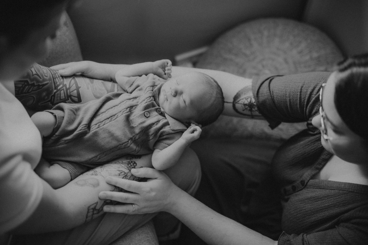 Black and white image of two parents cradling their newborn baby together on a sofa.