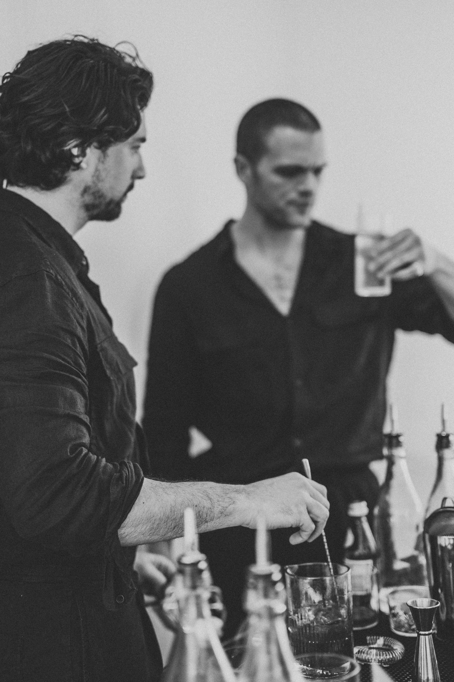 Two bartenders in black shirts preparing cocktails at a bar in black and white photography.