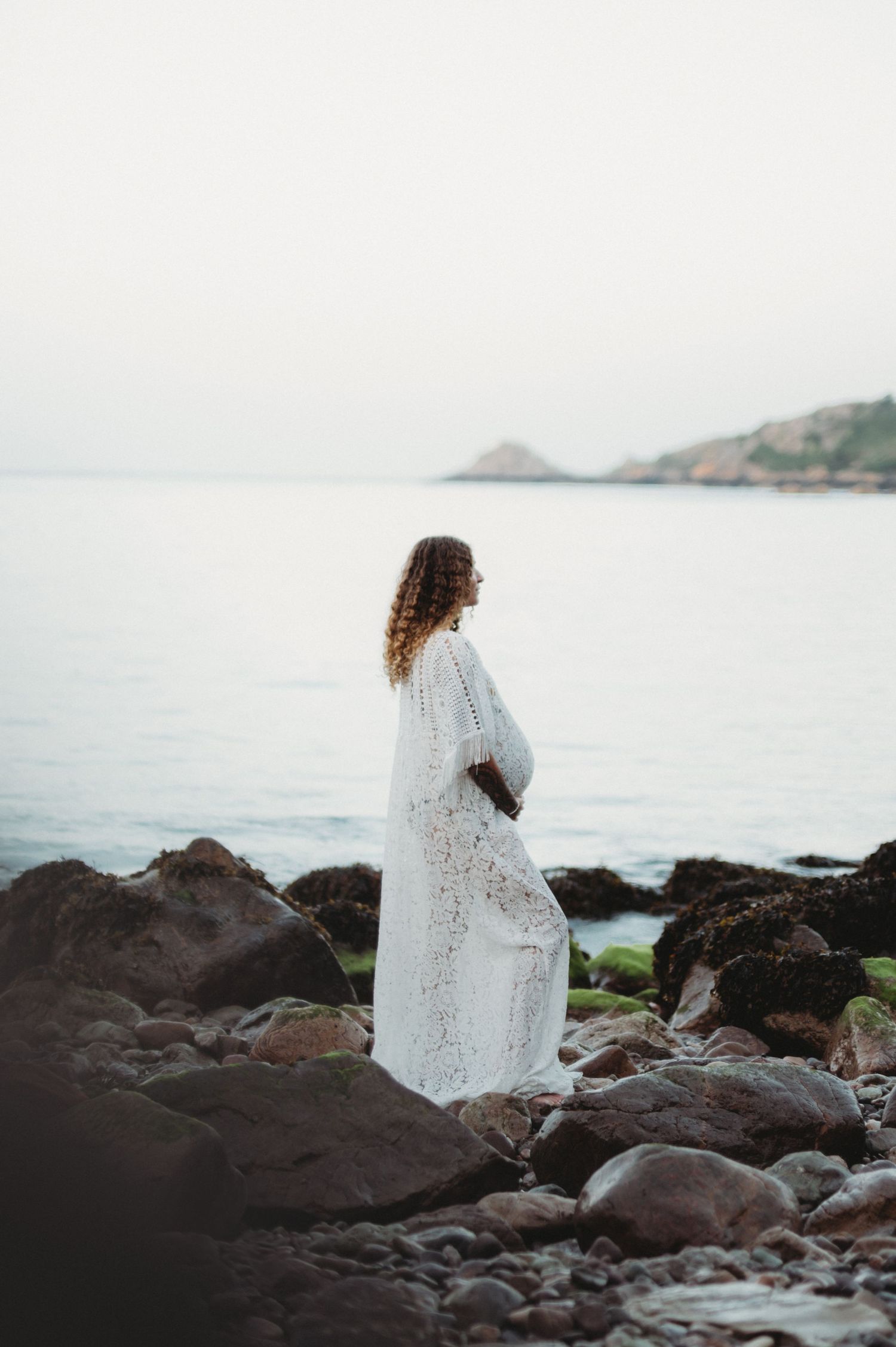 Pregnant woman in white lace dress standing on rocky beach shore gazing at calm ocean with misty hills in background.