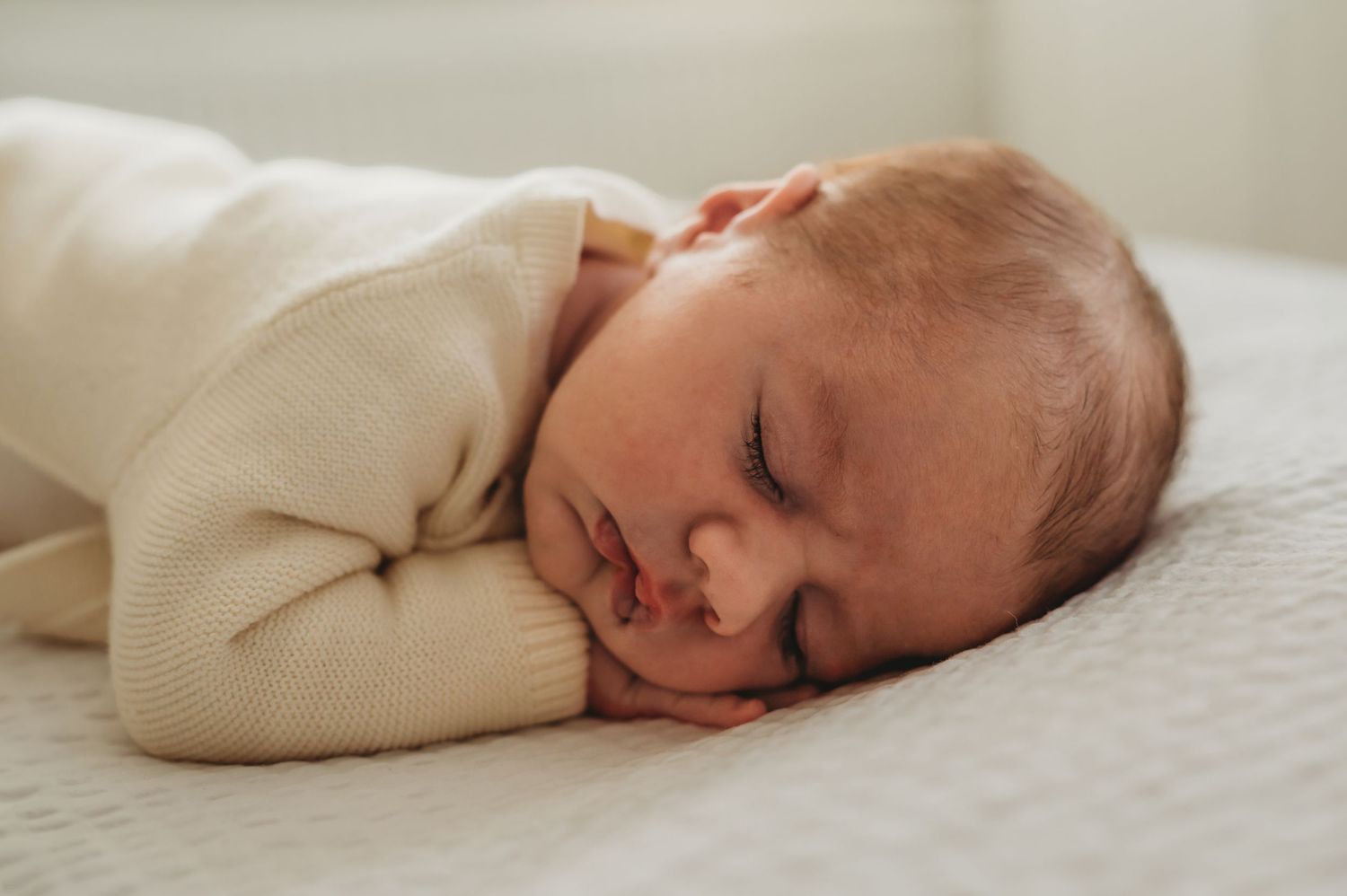A peaceful newborn baby sleeping soundly in a cream knit outfit on a white surface.