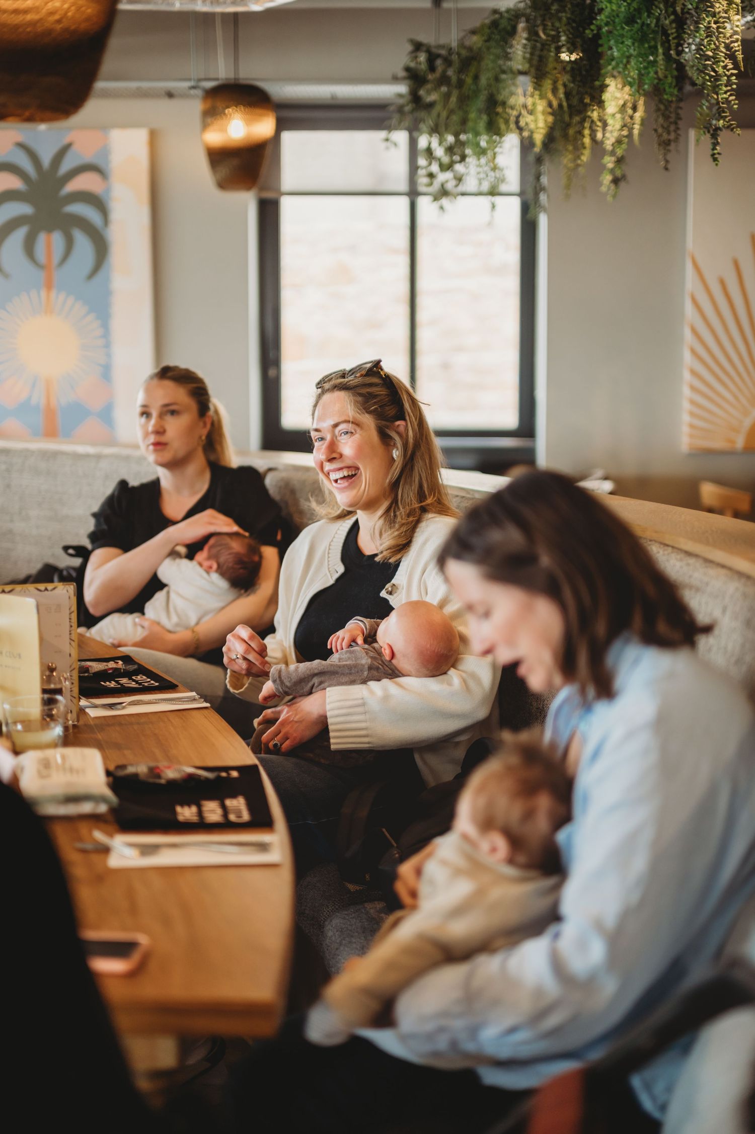 Three mothers breastfeeding newborns while socializing in a warm cafe setting.