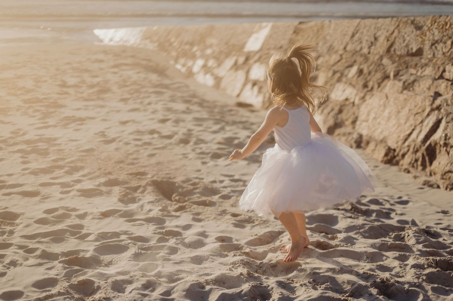Young girl in white tutu dress running barefoot on sandy beach at sunset.