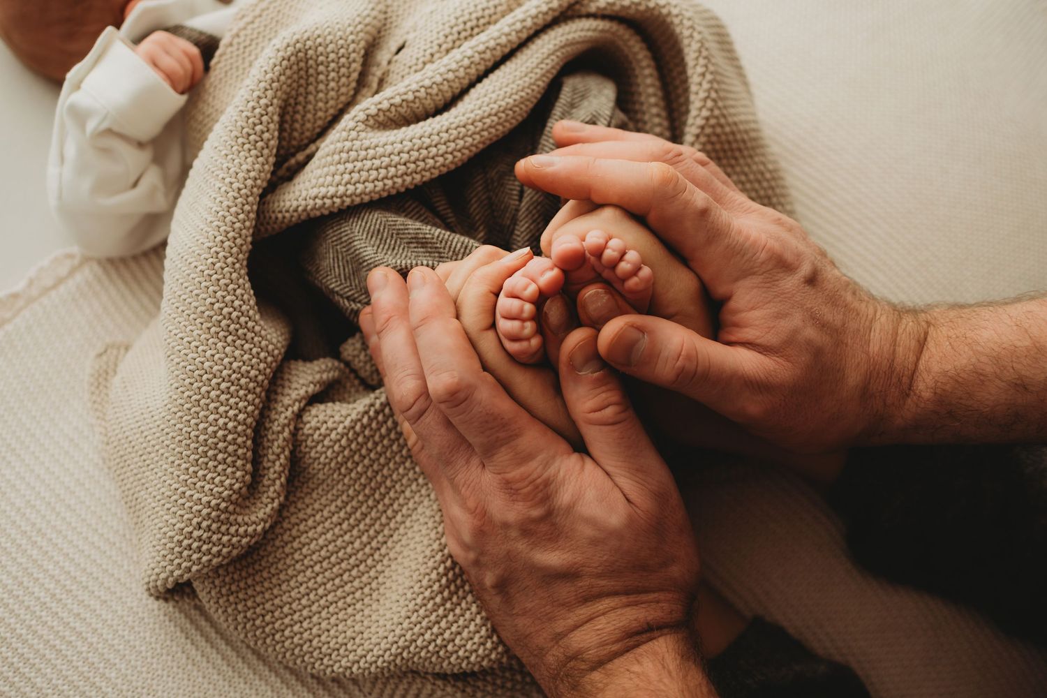 Adult hands gently cradling tiny newborn baby feet wrapped in a soft beige knit blanket.