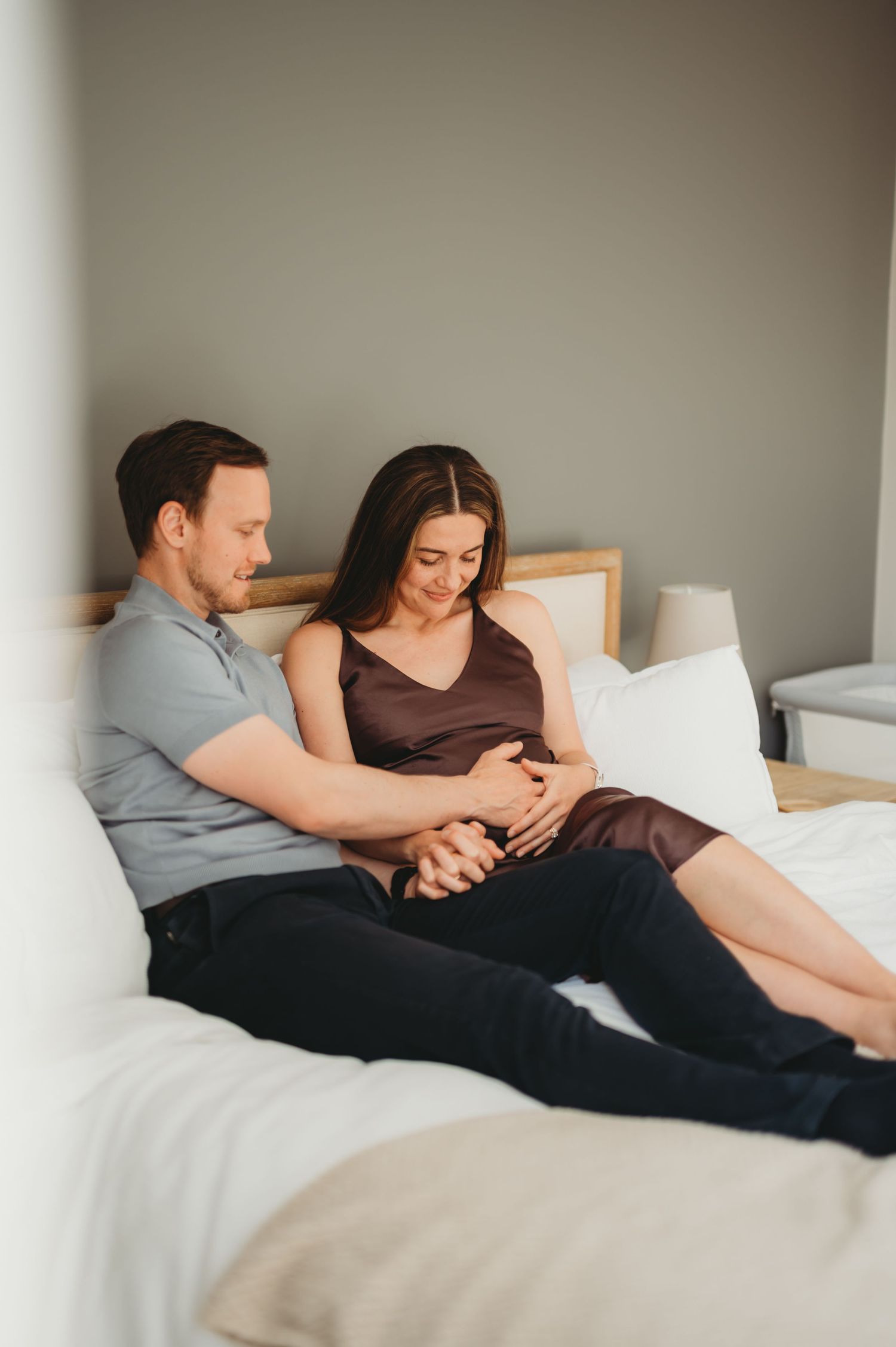 Couple relaxes on bed together, gently holding pregnant belly in a cozy bedroom setting.
