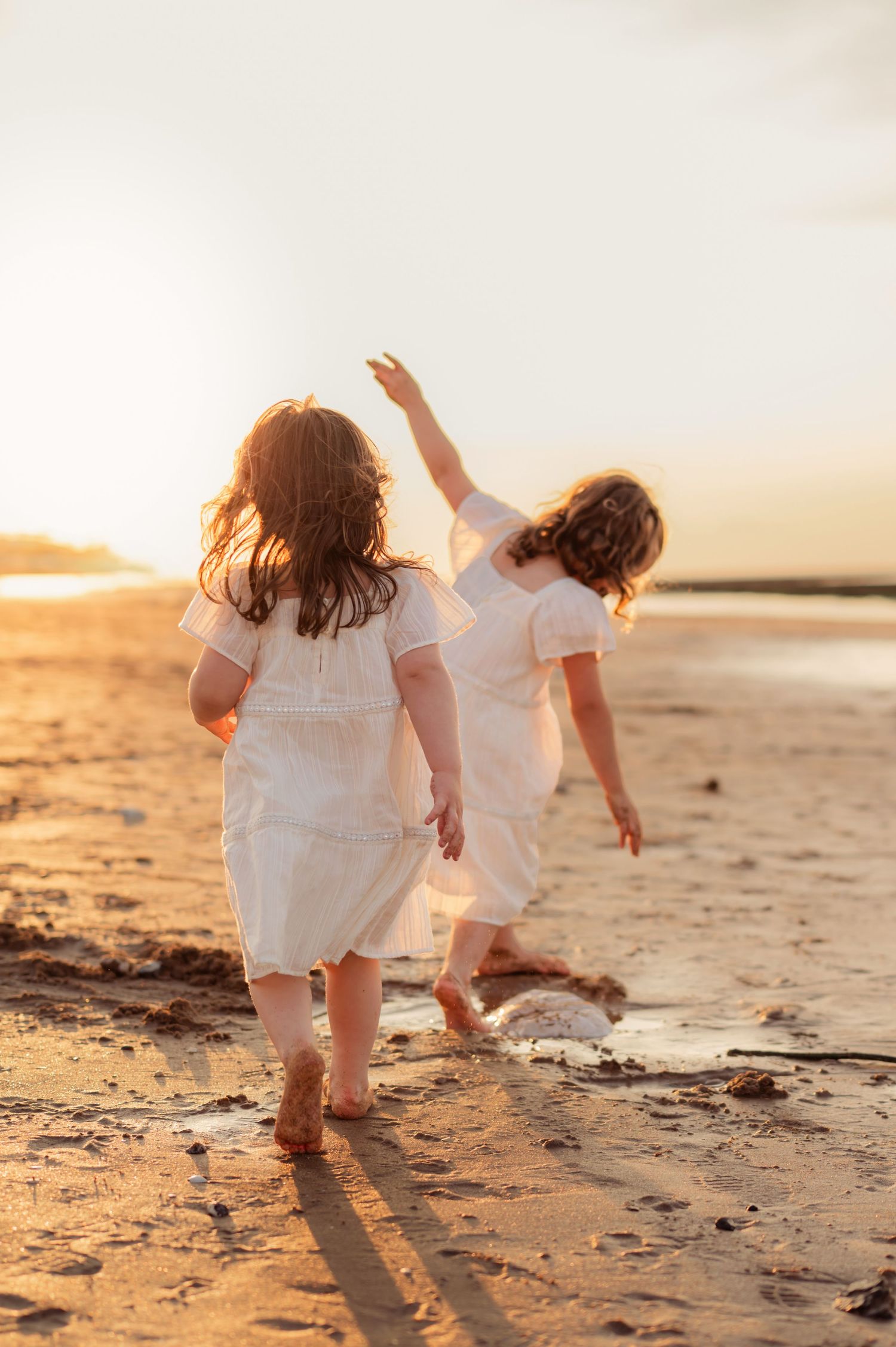 Two girls in white dresses walking barefoot on a sandy beach during a warm golden sunset.