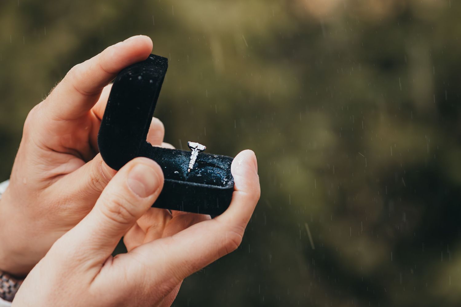 Hands holding an open black velvet box with a diamond engagement ring inside outdoors.