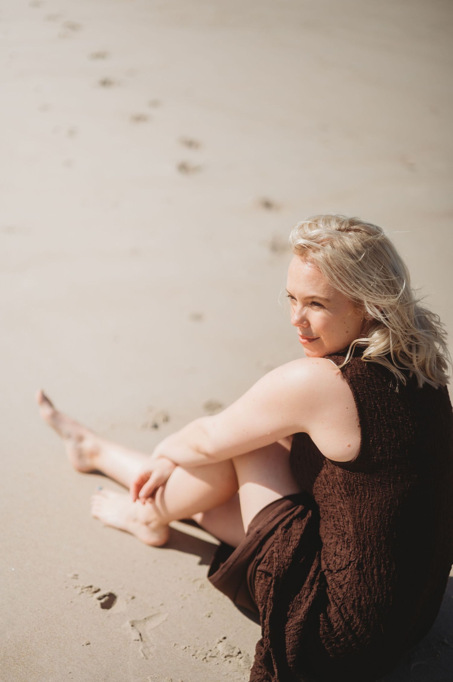 Blonde woman in brown dress sitting on sandy beach with footprints behind her.