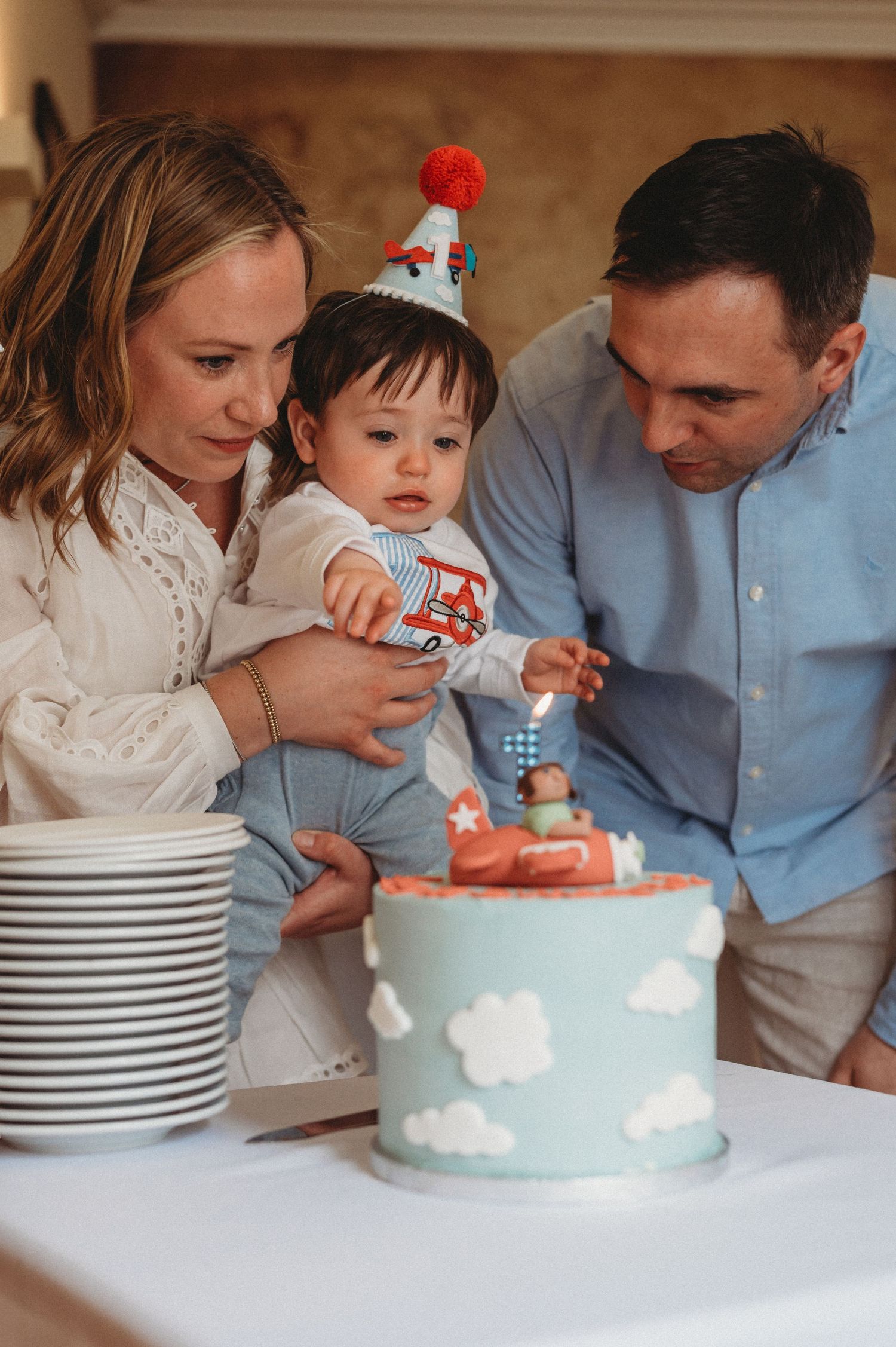 Parents and baby celebrating first birthday with a sky-themed cake decorated with clouds and an airplane.