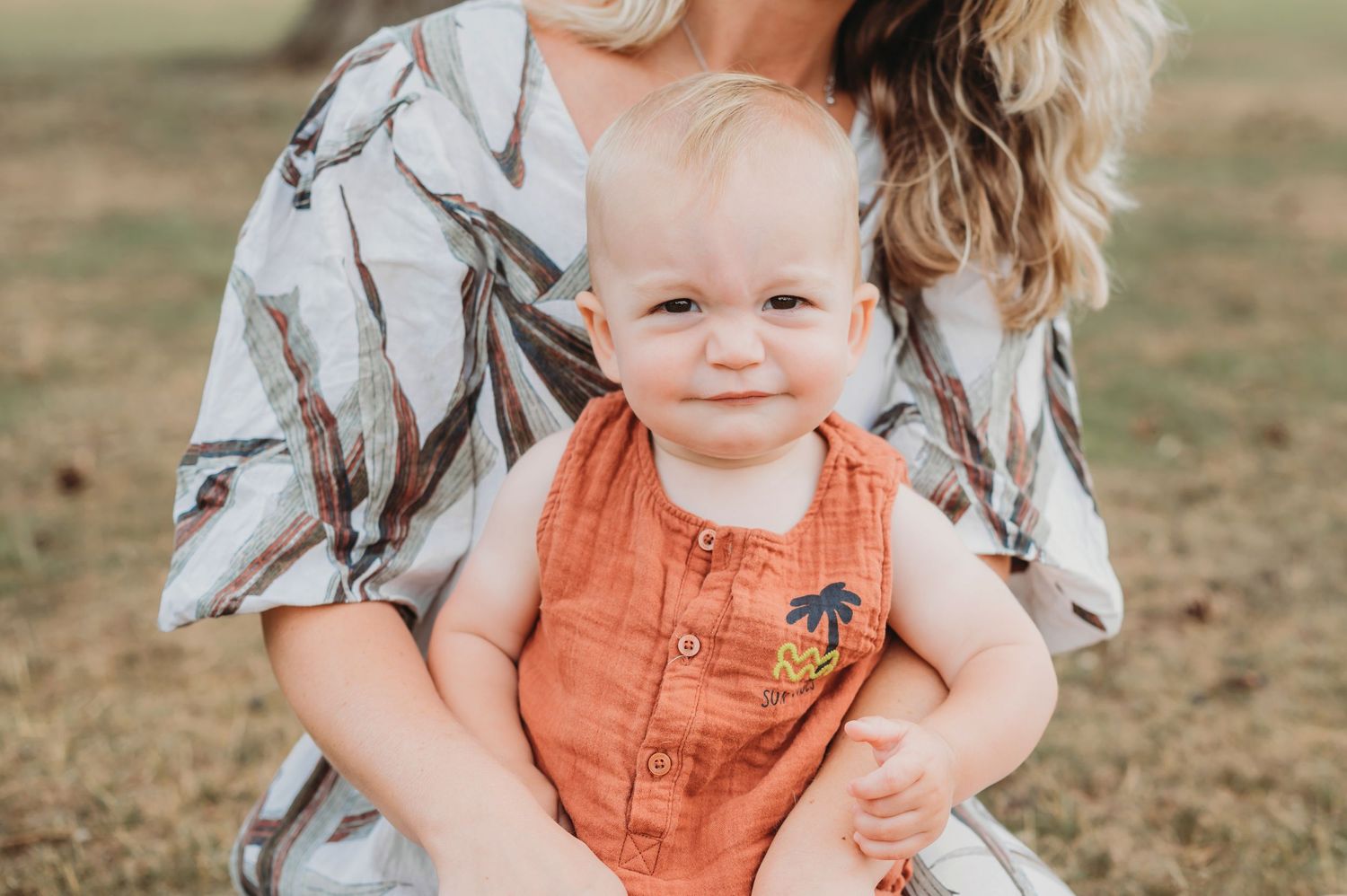 A smiling baby boy in an orange romper is held by his mother outdoors in a sunny field.