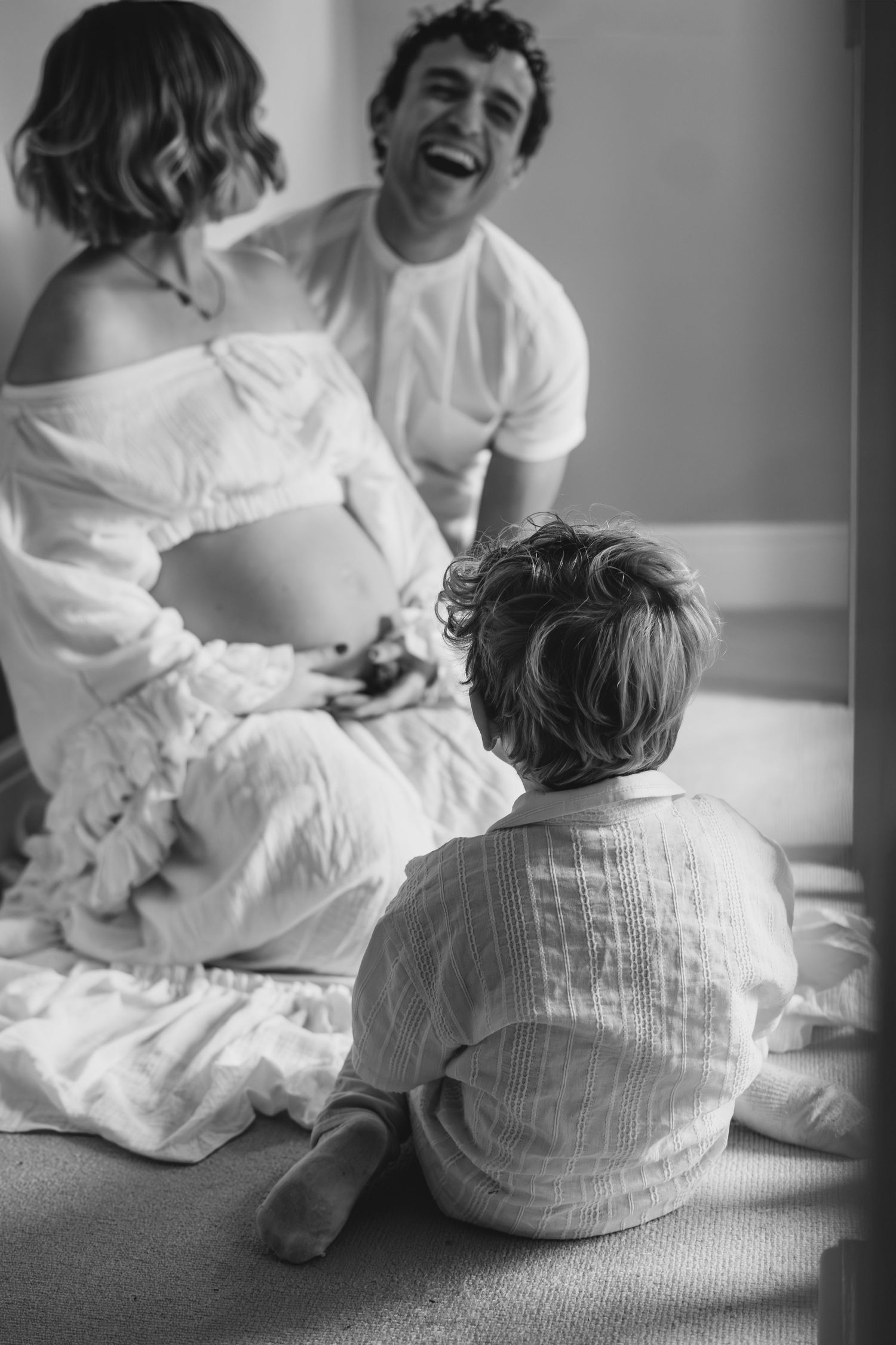 Black and white photo of laughing couple on bed with toddler sitting on floor before them.