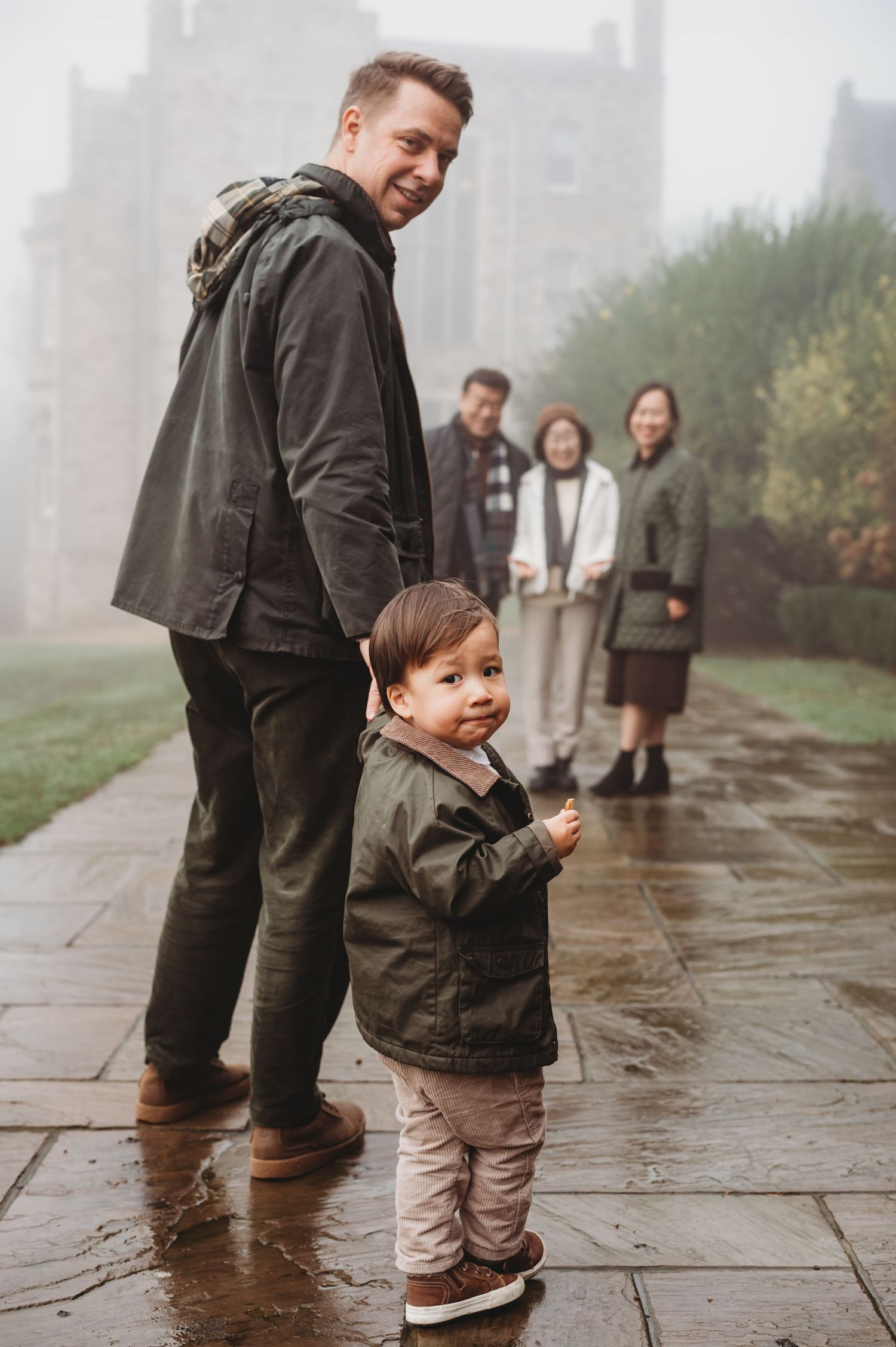 Smiling father and toddler in matching green jackets pose on a rainy foggy path with family behind.