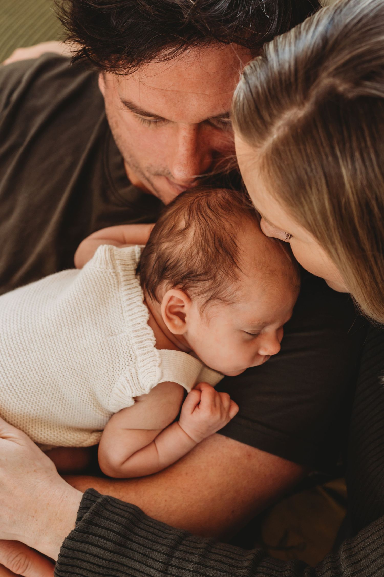 Loving parents gently lean in to kiss their sleeping newborn baby wrapped in a white knit blanket.