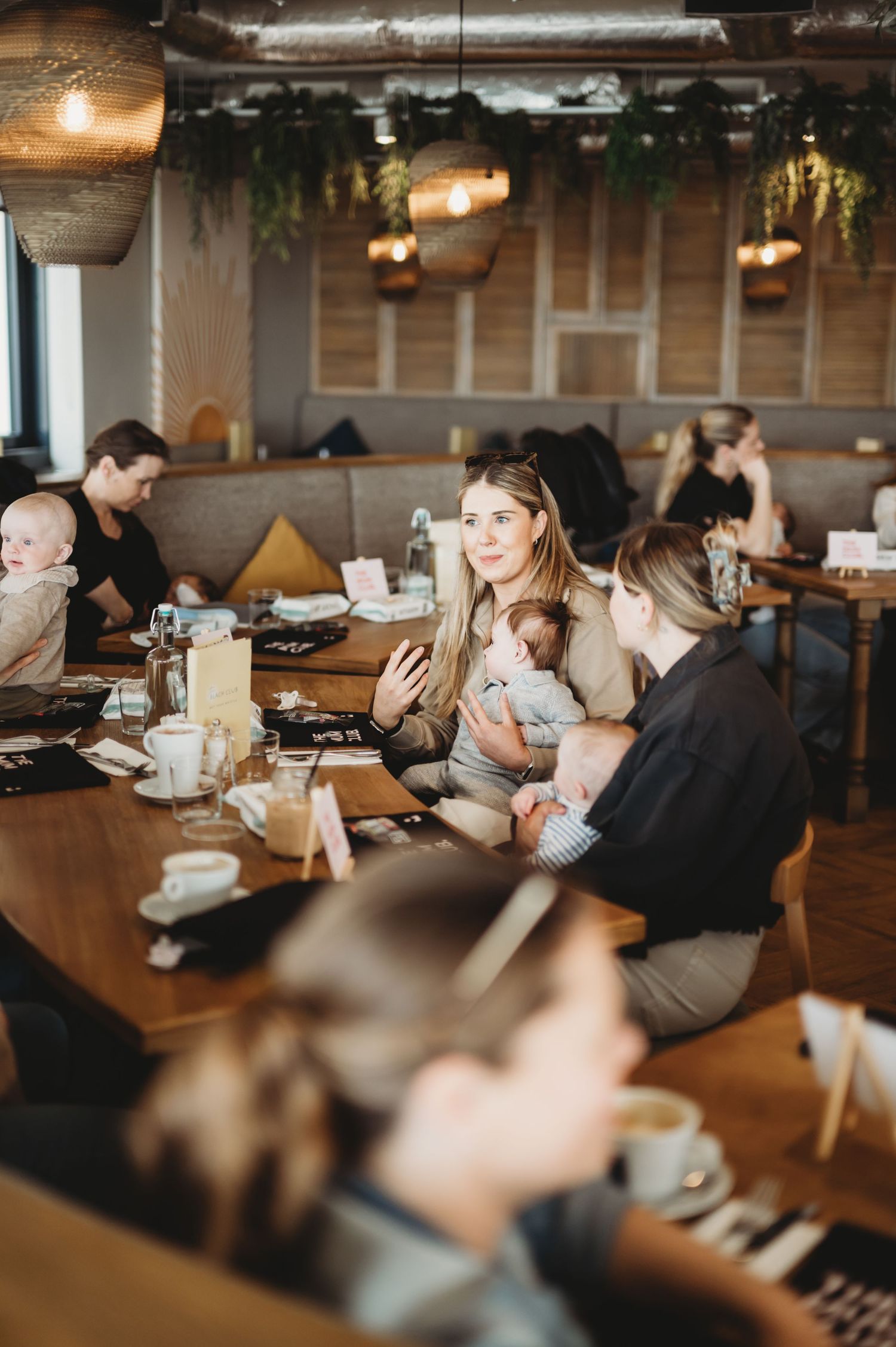Mothers with babies socializing at a cafe gathering around a wooden table with coffee cups.