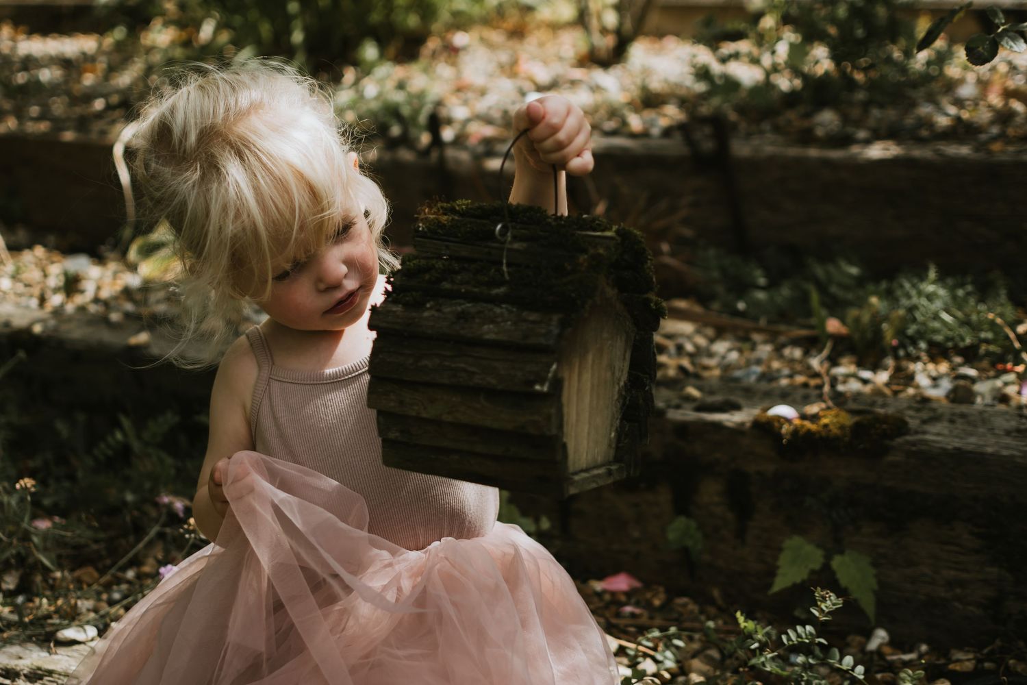 A young blonde toddler in a pink tutu dress explores a rustic wooden birdhouse in a garden setting.