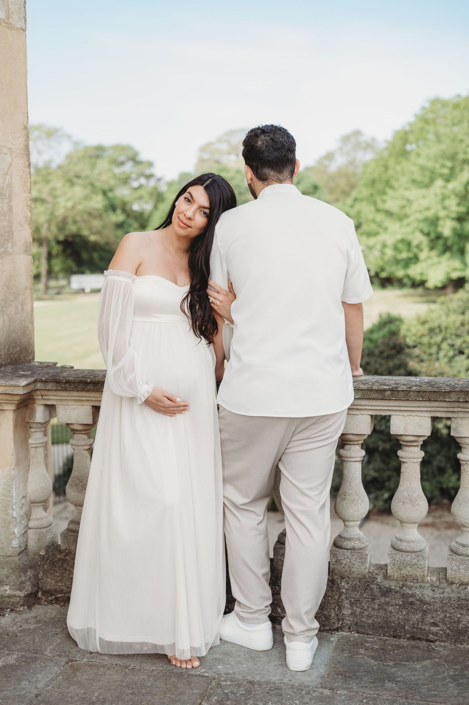 Elegant pregnant woman in white gown stands beside partner on stone balcony overlooking green park.