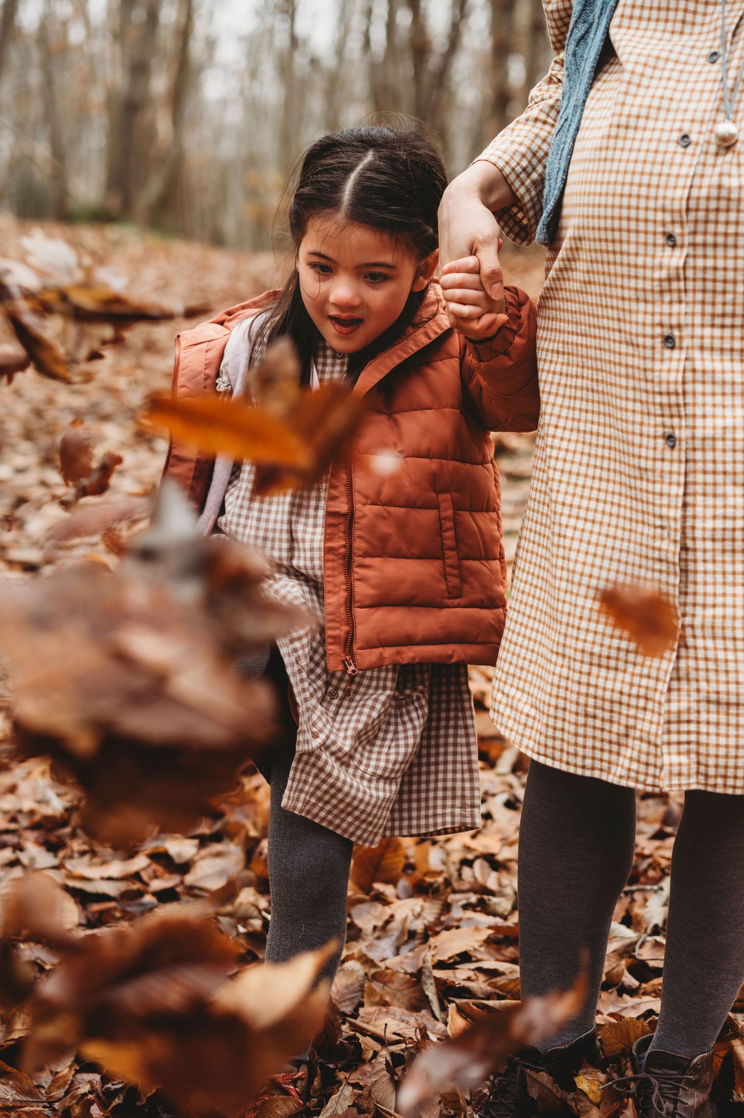 Young girl in an orange vest playing with falling autumn leaves in a forest.