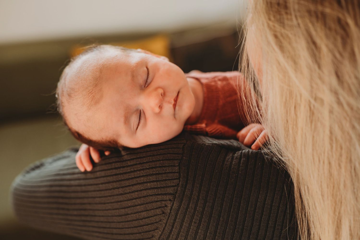 Sleeping newborn baby rests peacefully on a mother's shoulder wearing a rust-colored knit outfit.