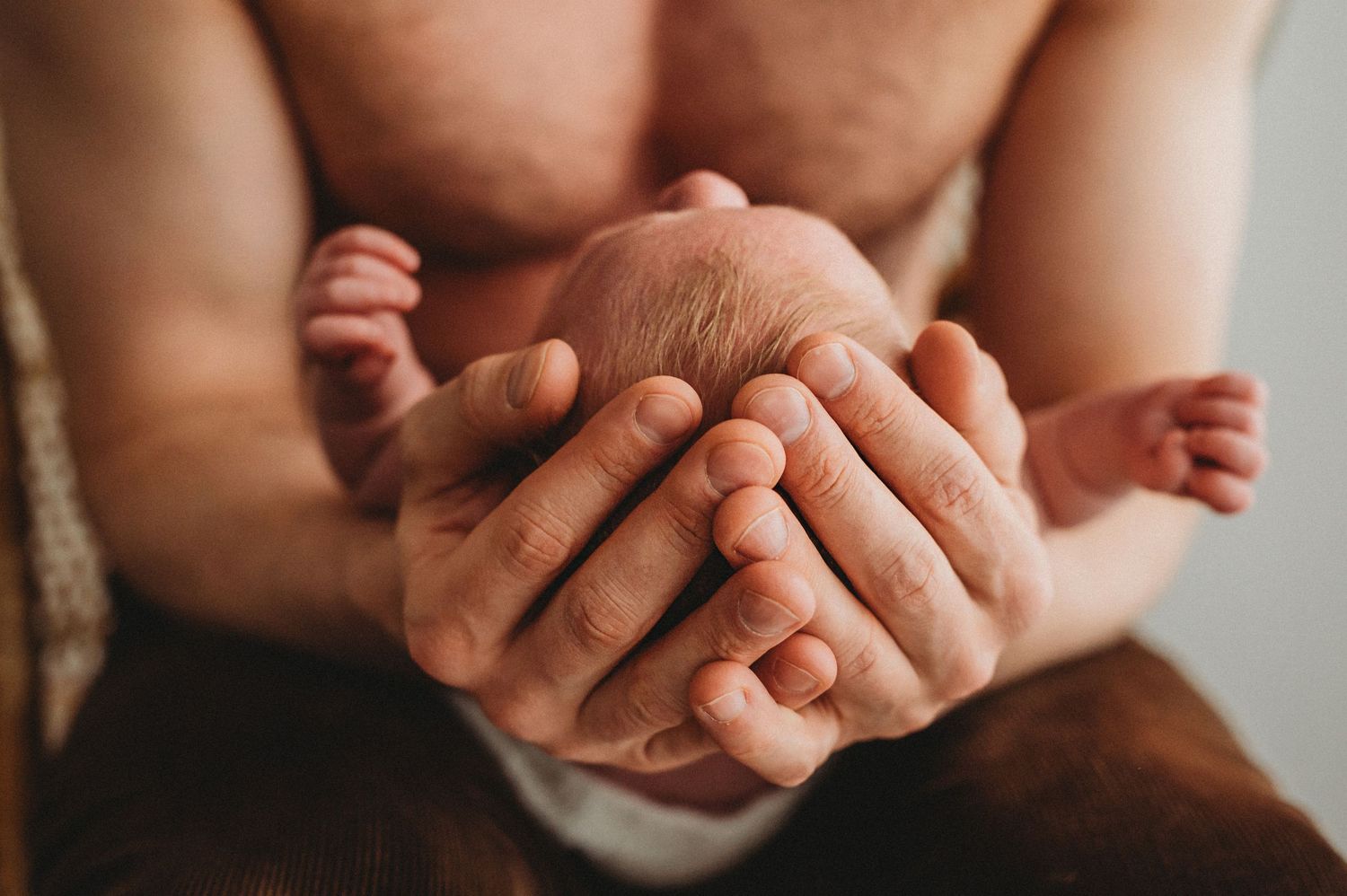 Father gently cradles sleeping newborn baby in both hands against his bare chest.