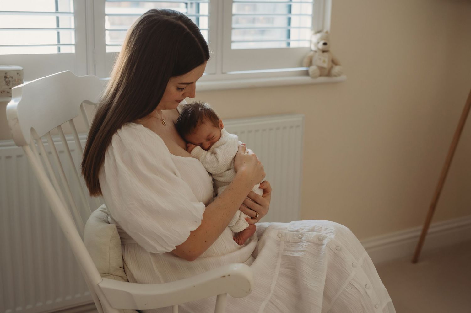 Mother in white dress sitting in rocking chair cradling sleeping newborn baby.