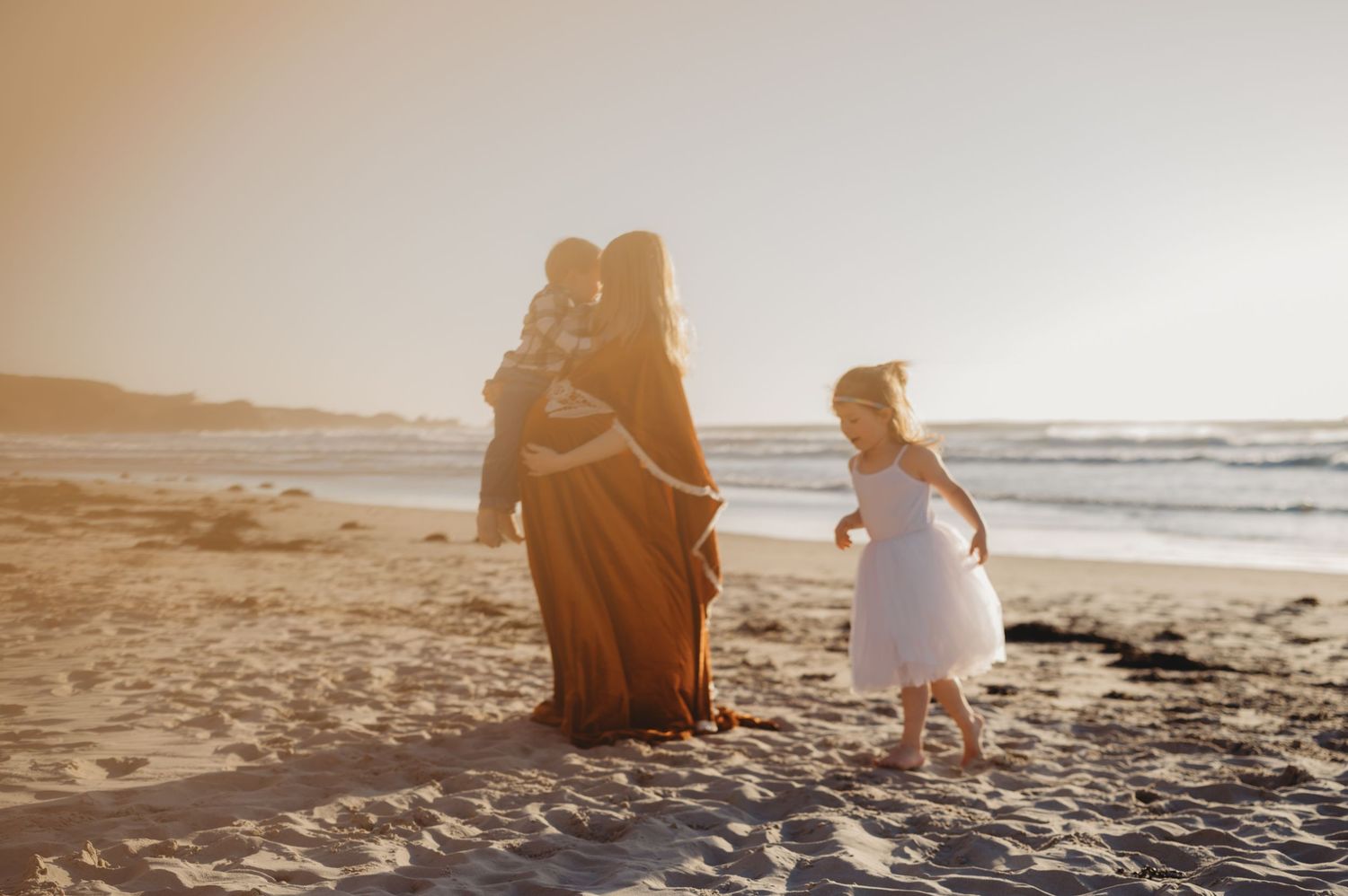 Mother in rust boho dress holding child at golden hour beach sunset with young girl running nearby.