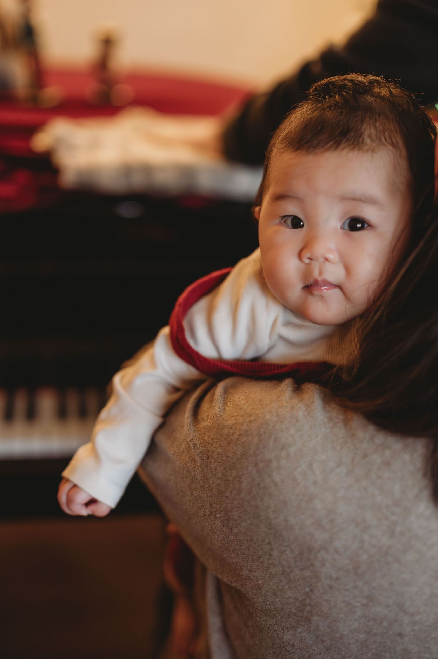 Adorable Asian baby in white outfit being held over a parent's shoulder near a piano.