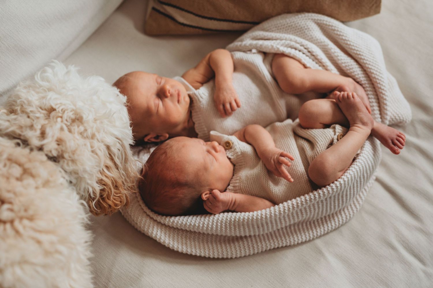 Two sleeping newborn twins in white onesies nestled together in a soft striped bassinet.