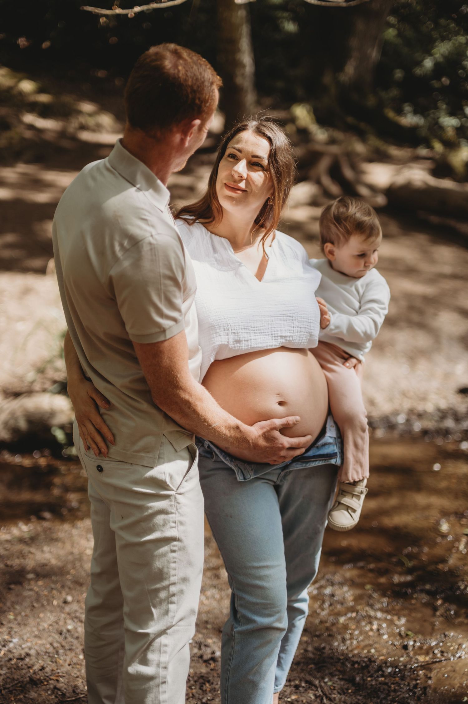 Pregnant woman with exposed baby bump stands with partner and toddler in a sunlit forest setting.