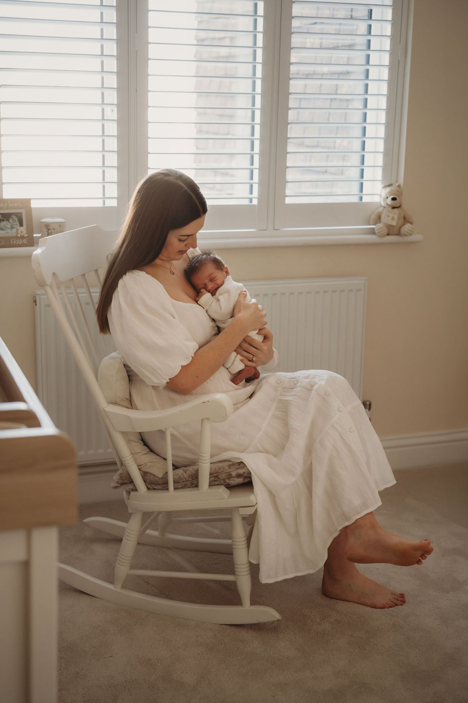 Mother in white dress sitting in rocking chair holding newborn baby by nursery window.