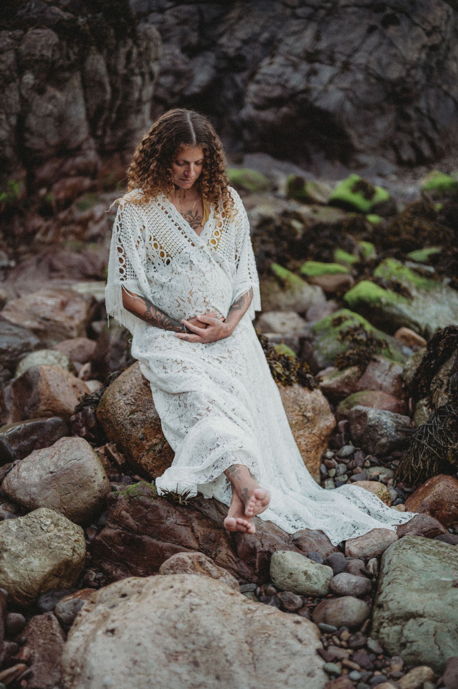 Pregnant woman in white lace dress sitting on rocky shore, cradling her belly in a serene maternity photo.