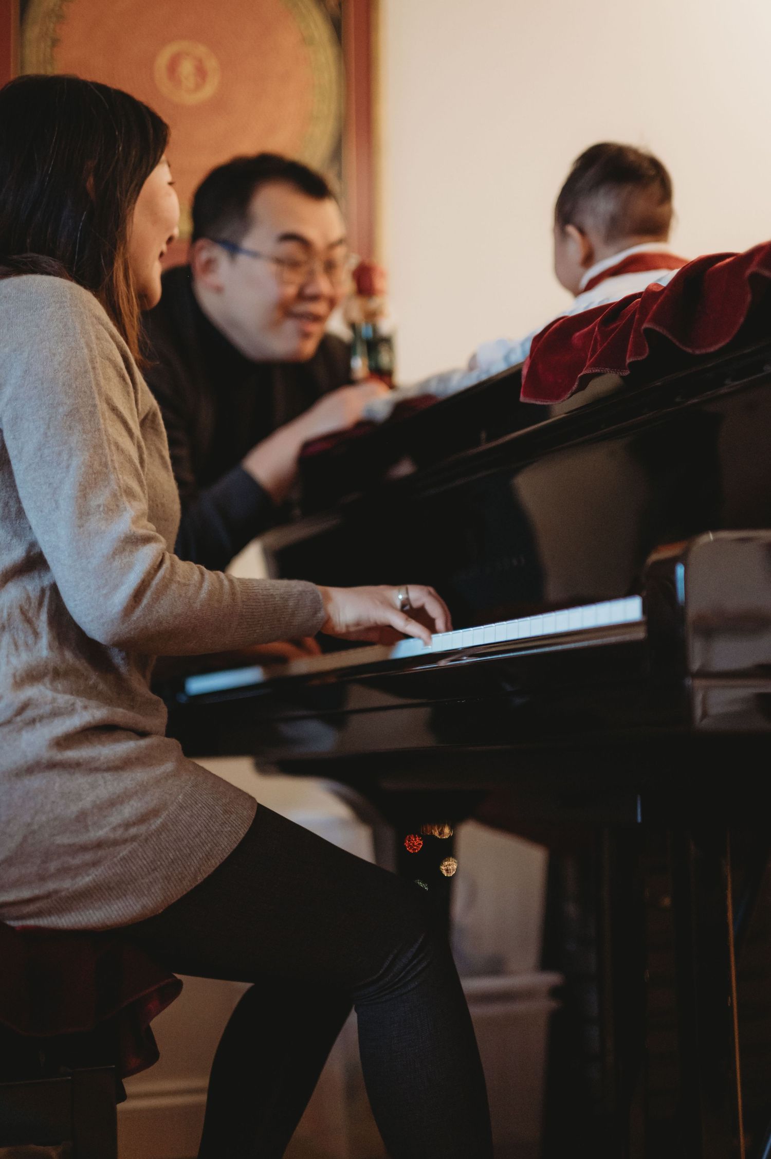 Asian woman playing a grand piano while a man smiles and a baby watches in a cozy room.