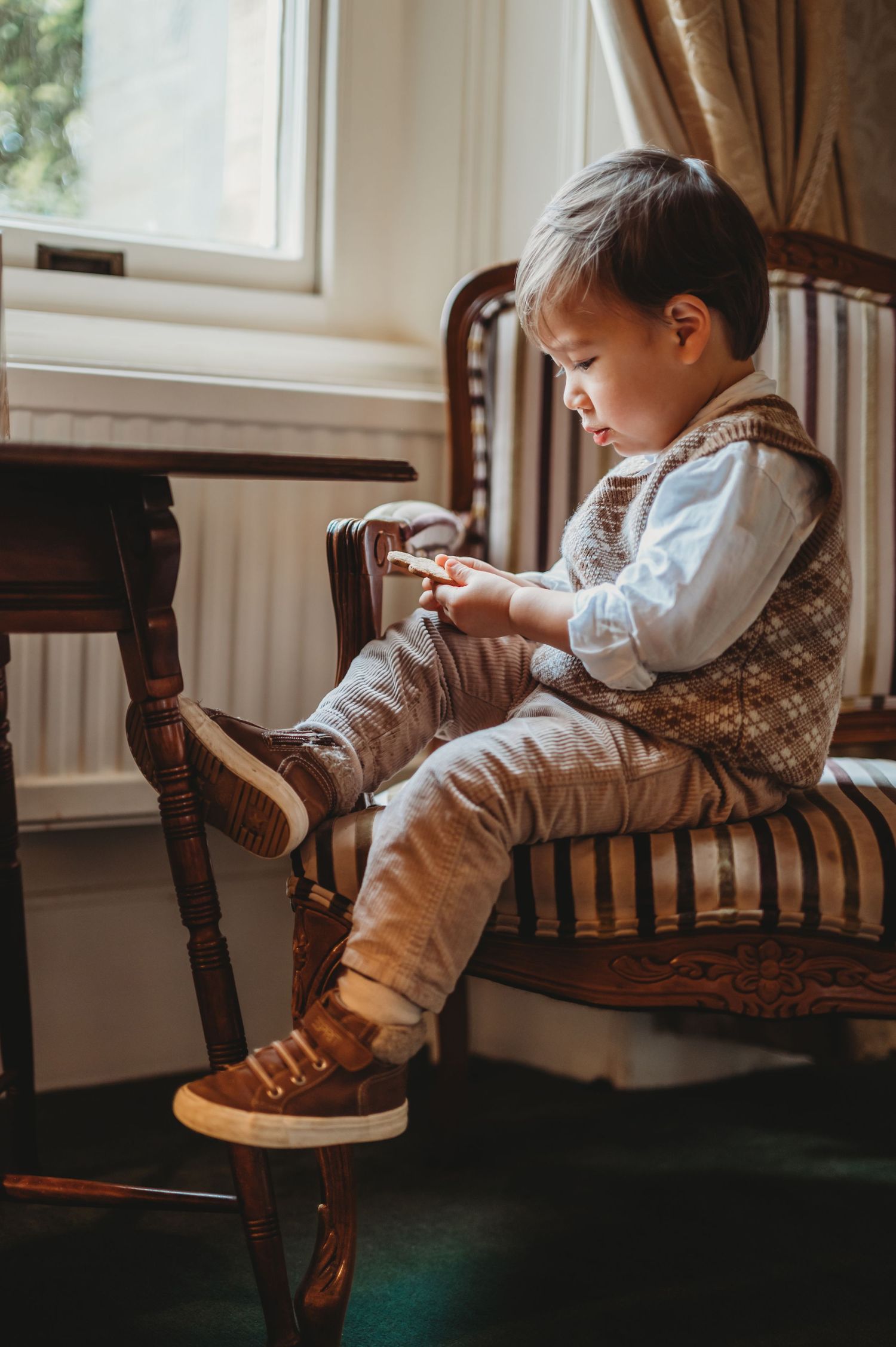 Young boy in a vest sitting on an antique chair by a window playing with a toy.