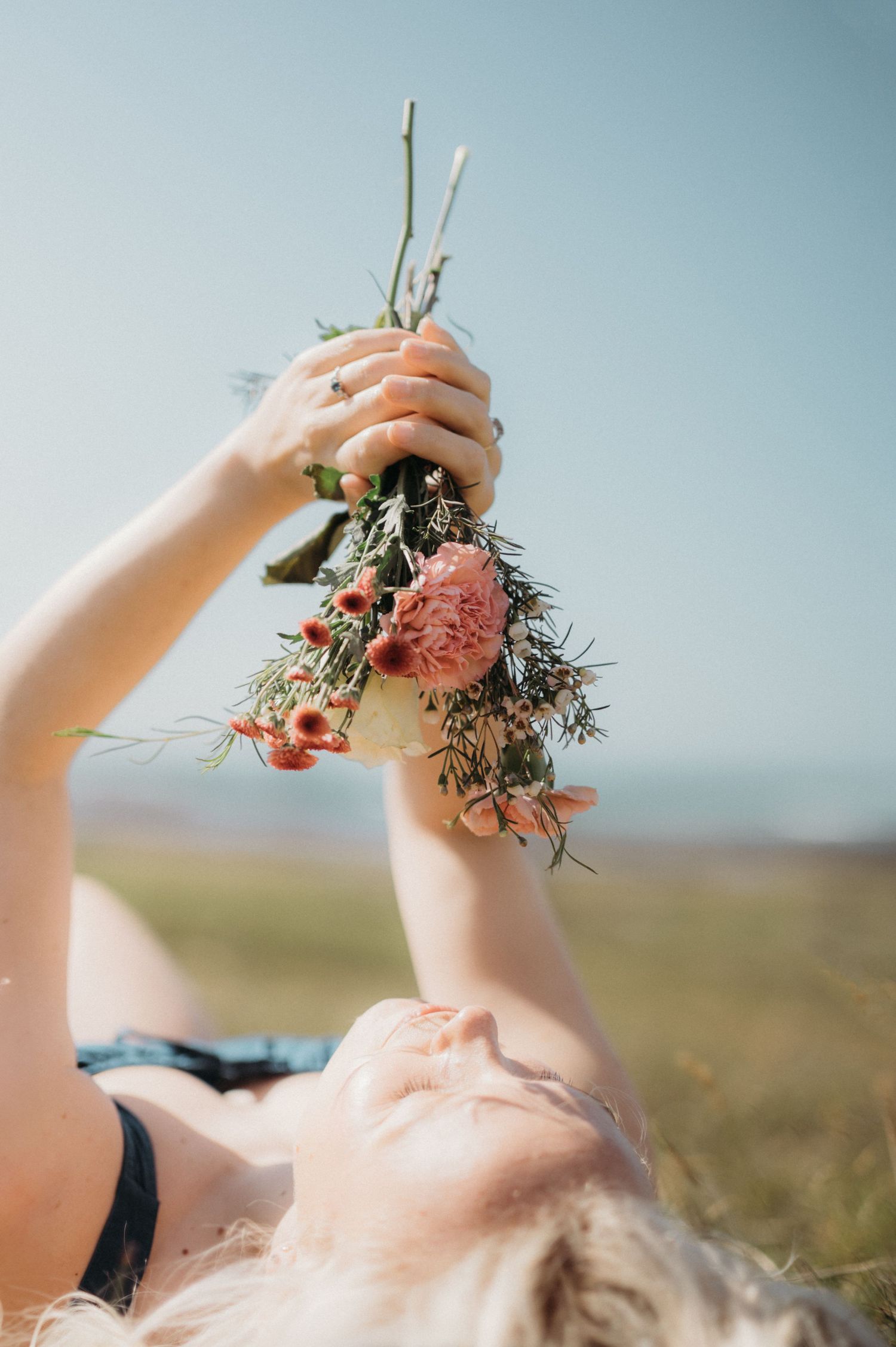 Blonde woman lying in field holding wildflower bouquet toward blue sky on sunny day.
