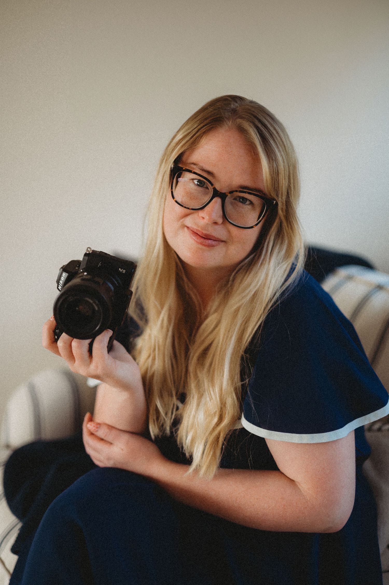 A blonde woman with glasses holds a camera while sitting on a couch.