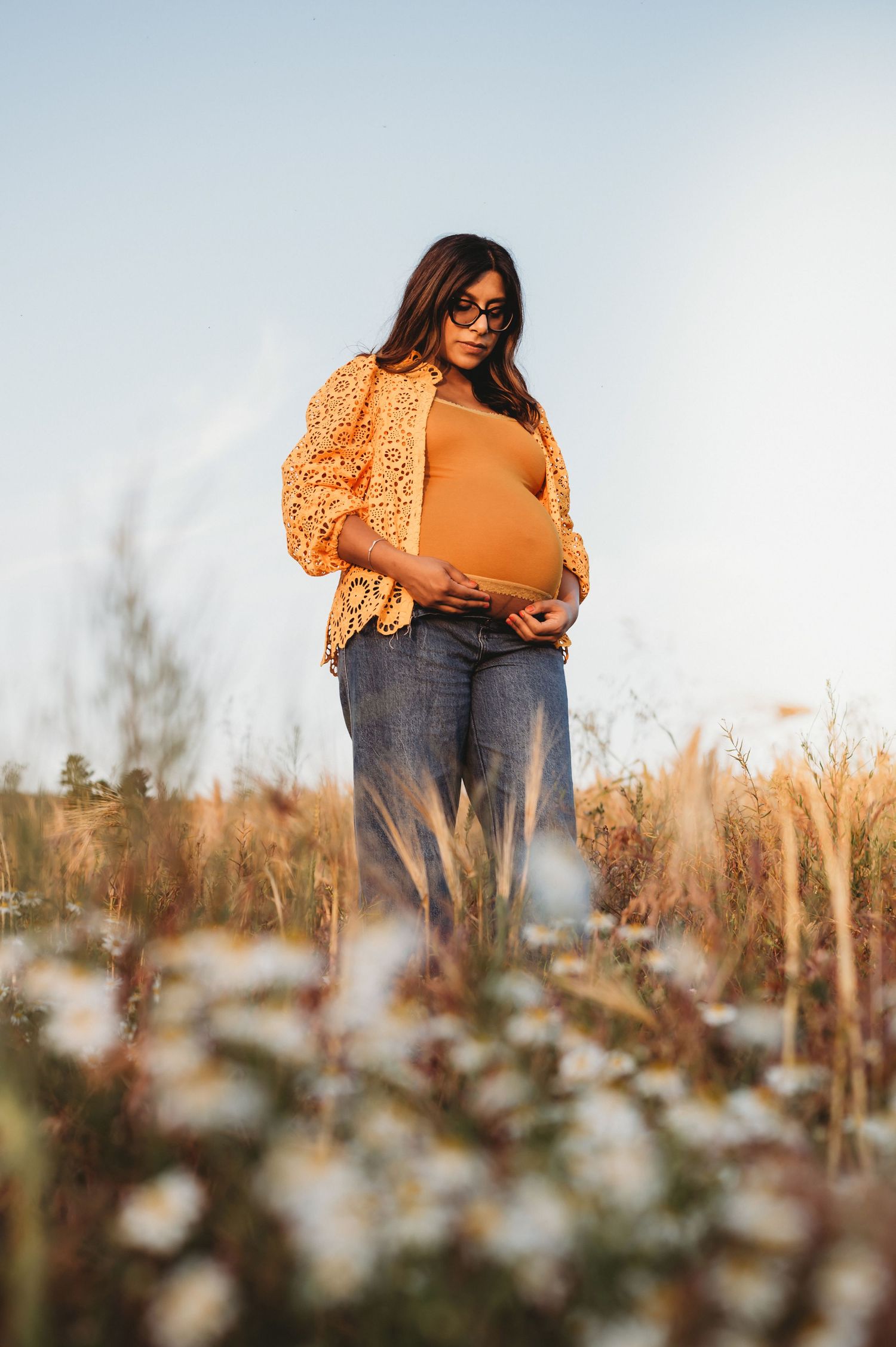 Pregnant woman in orange top and jeans stands in a wildflower field at golden hour.