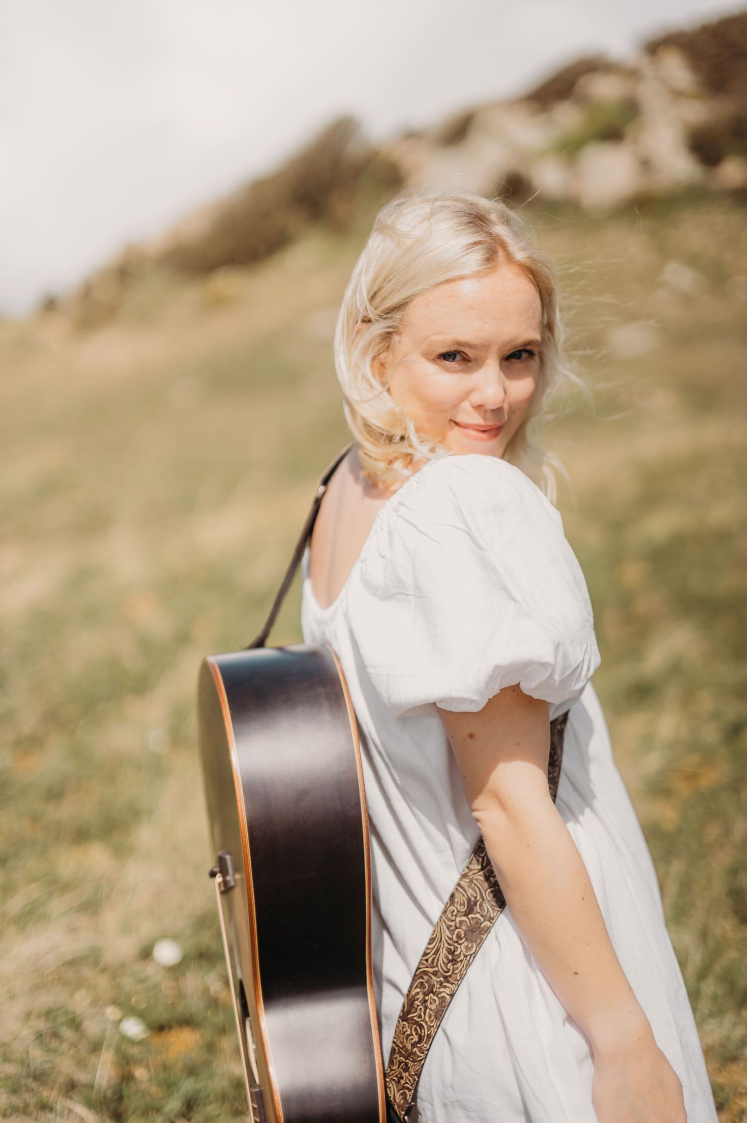 Blonde woman in white puff-sleeve dress carrying an acoustic guitar in a hillside field.