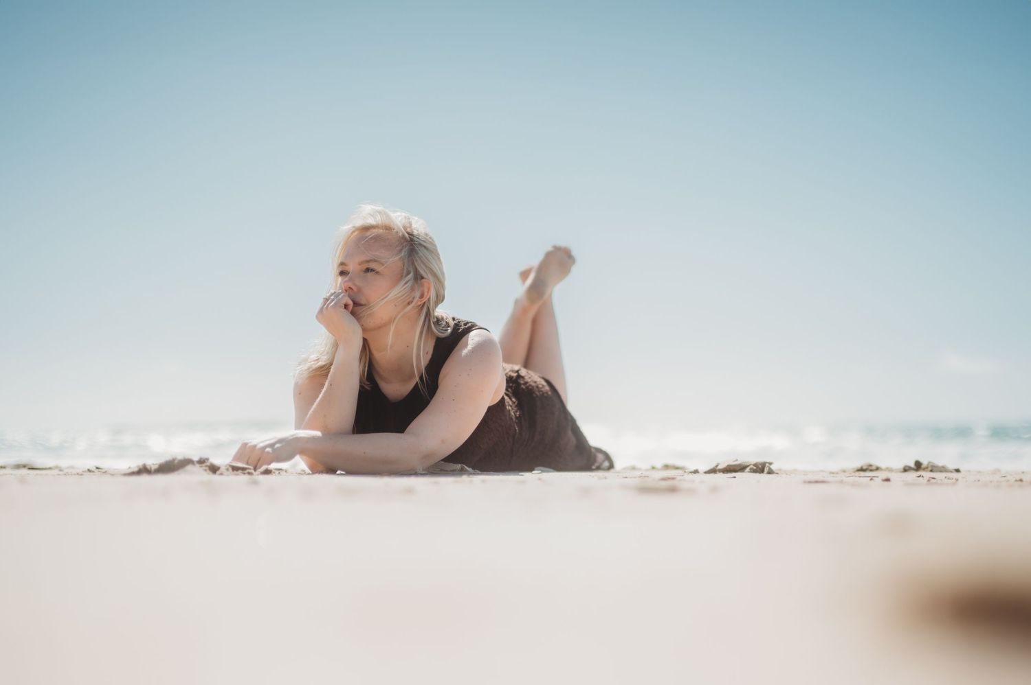 Blonde woman in black dress lying on sunny beach with ocean in background.
