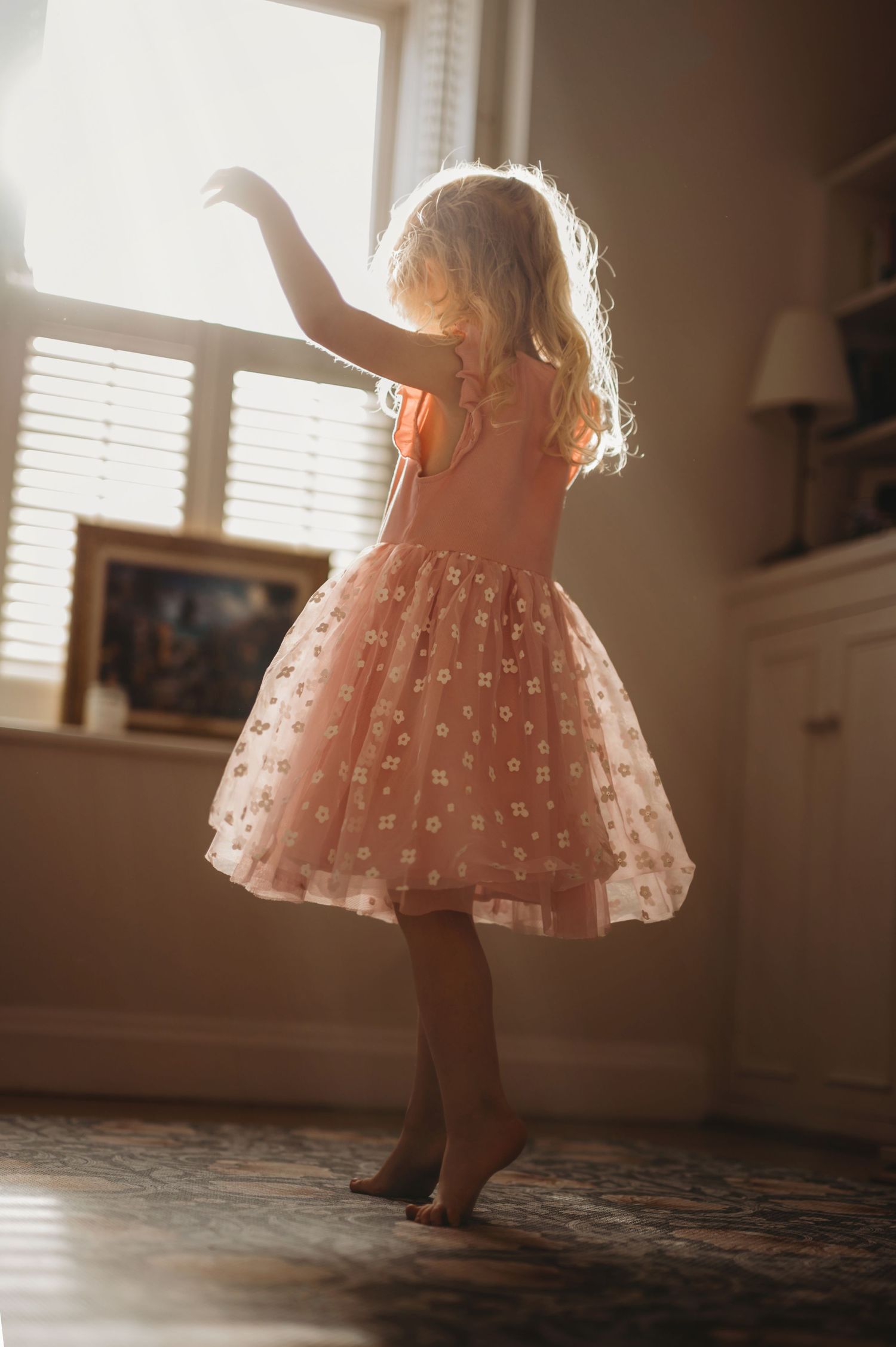 A young girl in a pink tulle dress twirls barefoot in a sunlit room.