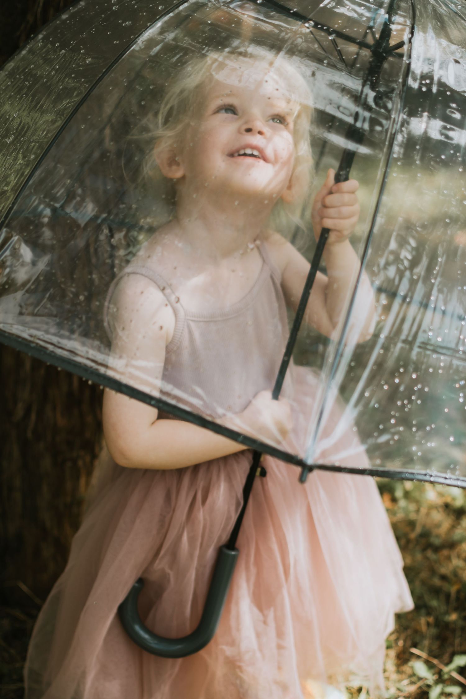 Joyful little girl in pink tutu dress smiling under clear bubble umbrella in rainy garden.