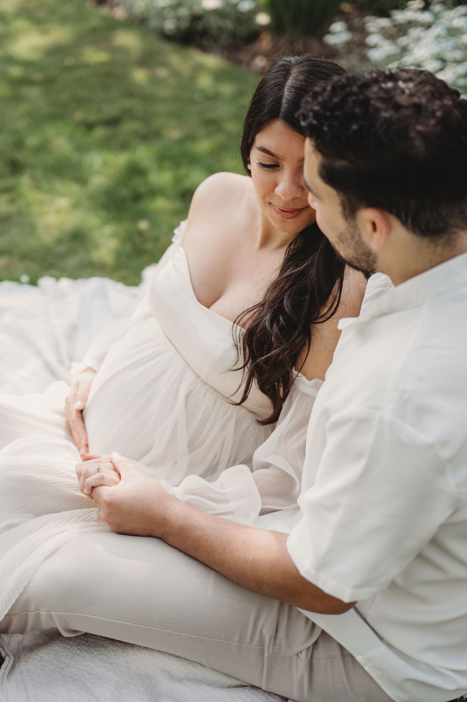 Couple sharing tender moment outdoors, pregnant woman in white off-shoulder dress holding hands with partner on blanket.