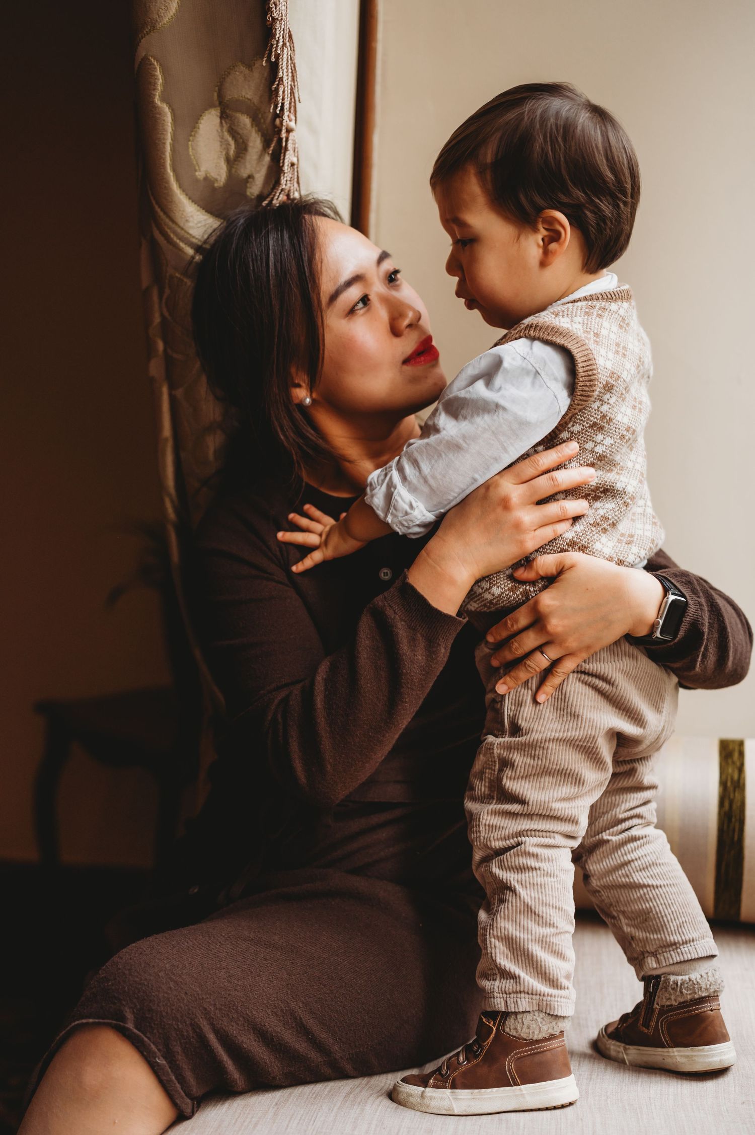 A mother sits on the floor holding her baby boy up while sharing a tender moment indoors.
