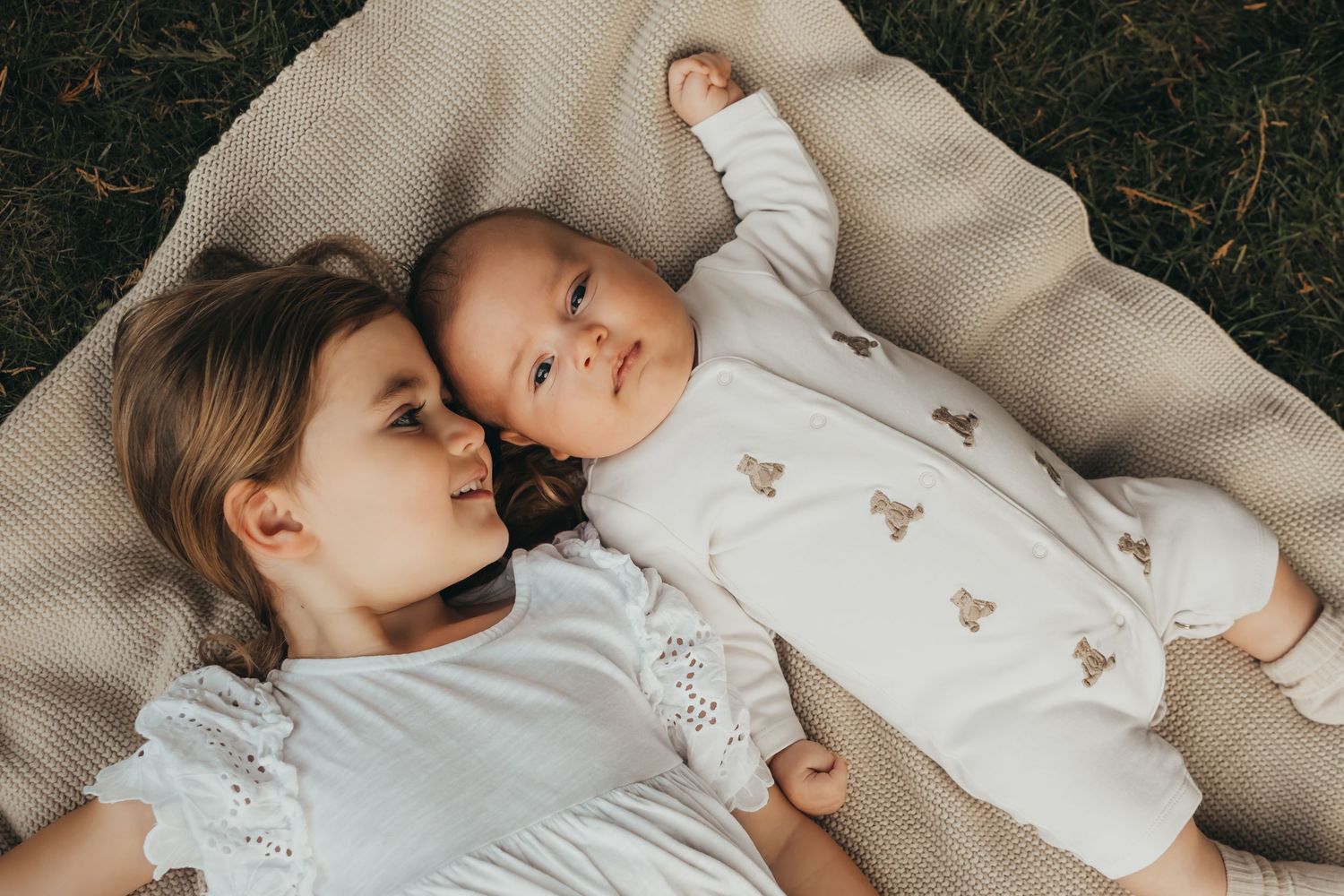 Young girl lying on a blanket outdoors cheek to cheek with an infant in a white bear onesie.