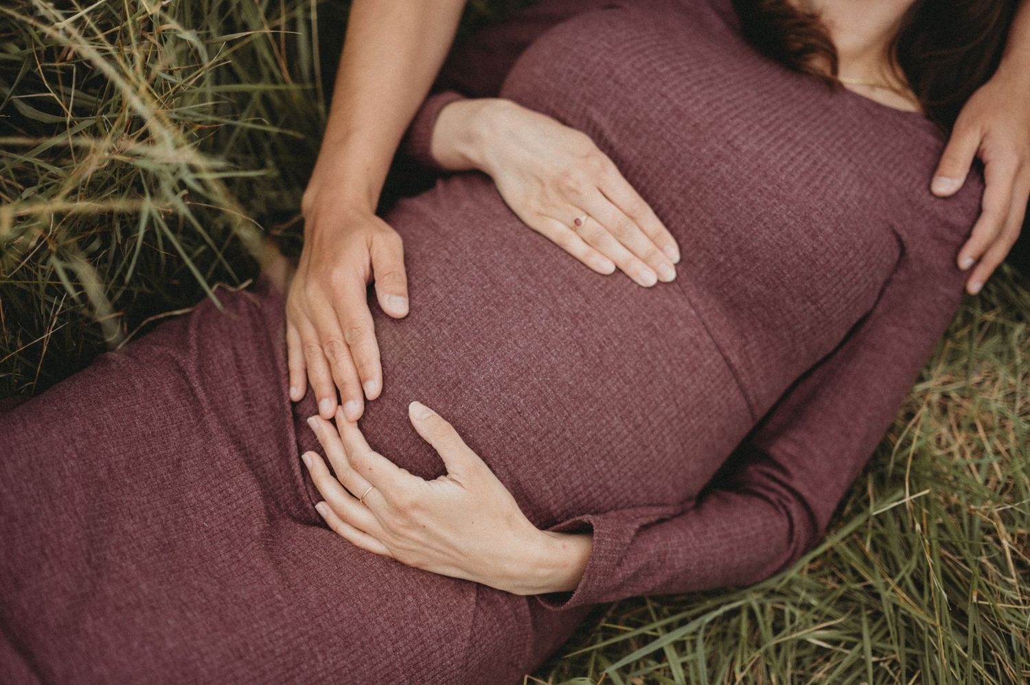 Close-up of couple's hands gently cradling a pregnant belly in a burgundy ribbed maternity dress.