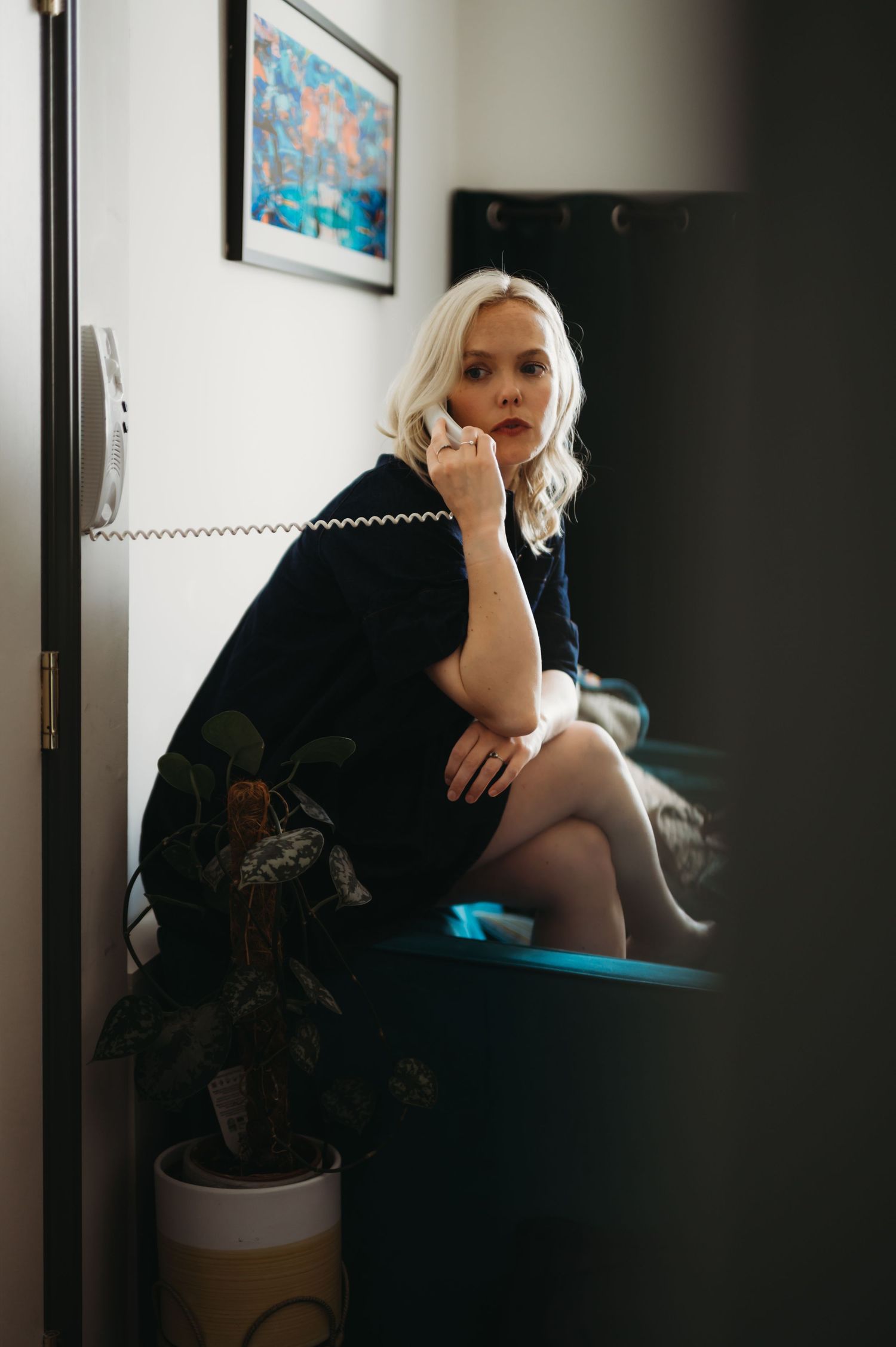 Blonde woman in black outfit sitting and talking on a retro wall phone indoors.