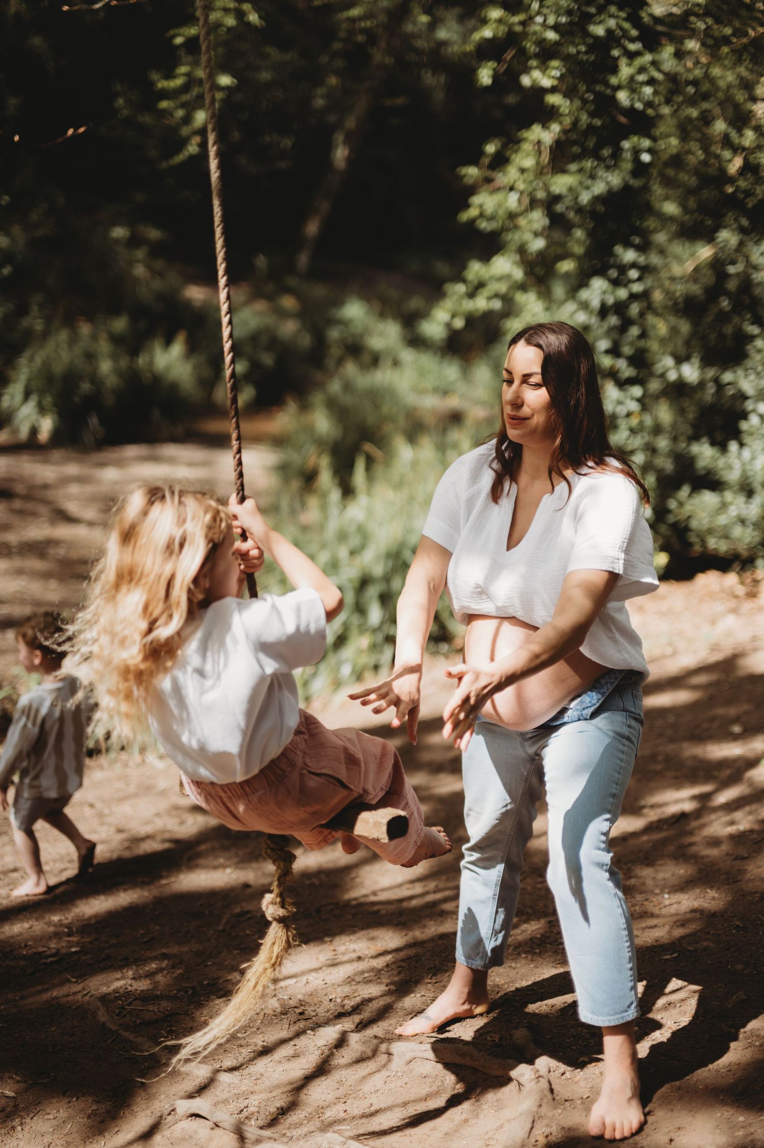 Pregnant woman pushing young girl on rope swing in sunny forest setting.