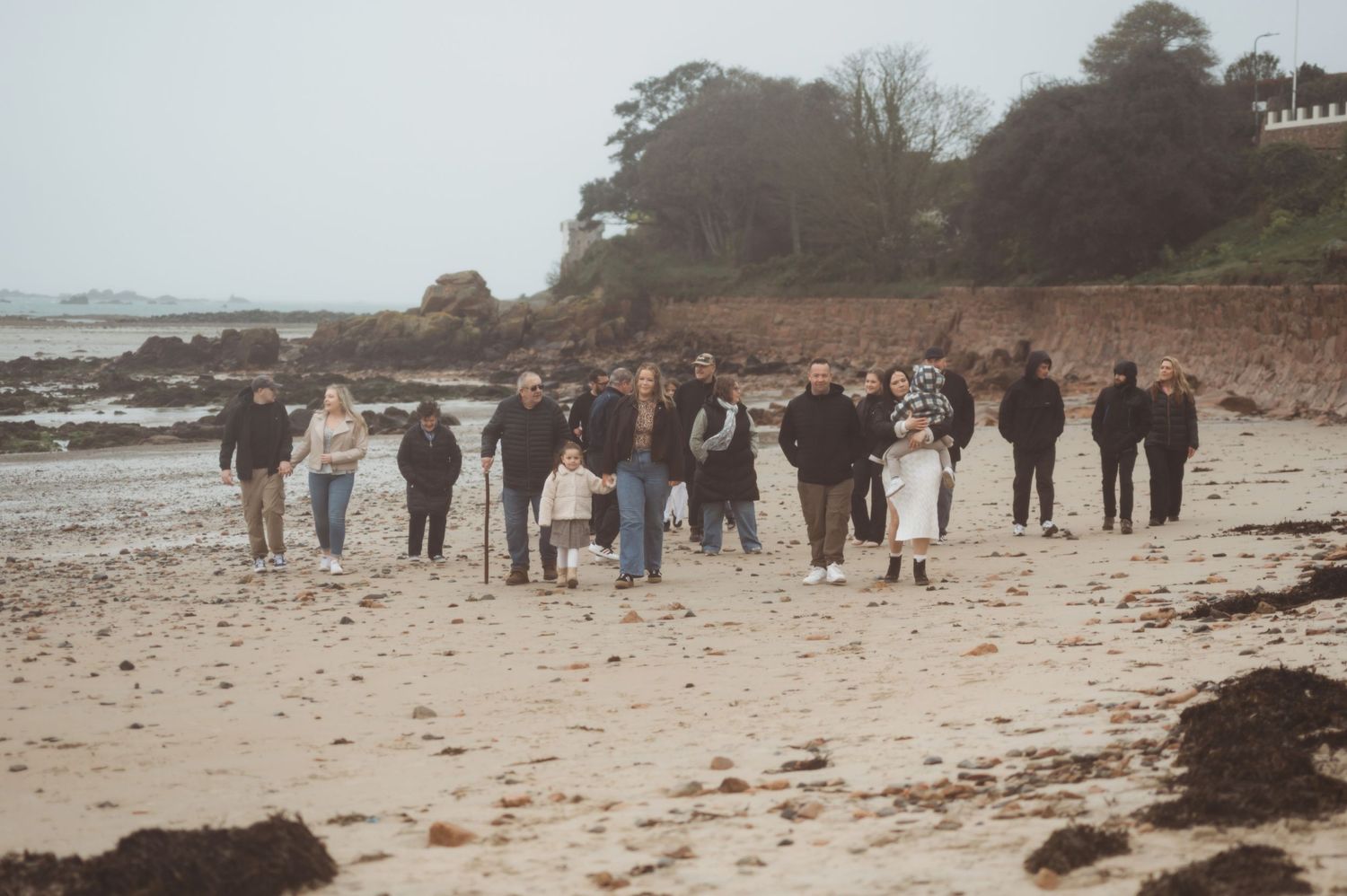 Group of people walking along a sandy beach on an overcast day near rocky coastline and trees.